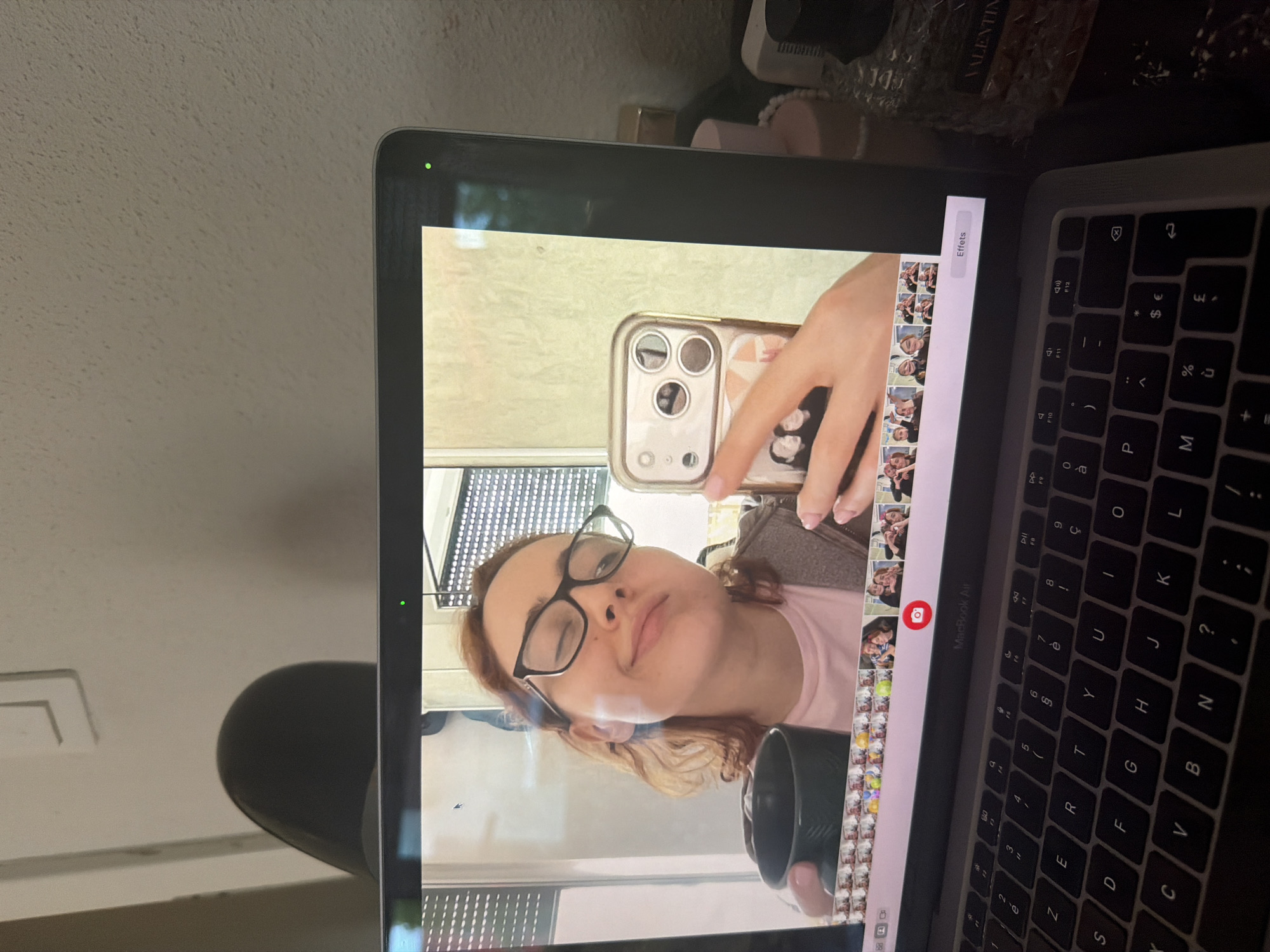 The image shows a woman taking a selfie in her laptop's camera. She is wearing glasses and a pink t-shirt. The laptop is open on a desk, and part of the keyboard is visible. In the background, there are some decorative items, including what appears to be a bottle of "VALENTIN" perfume. The scene is indoors, and the lighting suggests it could be daytime. The woman's expression is relaxed and playful. The laptop screen displays a row of thumbnails, indicating she might be using a photo editing application or browsing through pictures. A green light on the laptop suggests the camera is active. The overall context suggests a casual moment of self-documentation. The text "MacBook Air" and "Effets" is visible on the screen, indicating the laptop model and a feature related to photo effects.