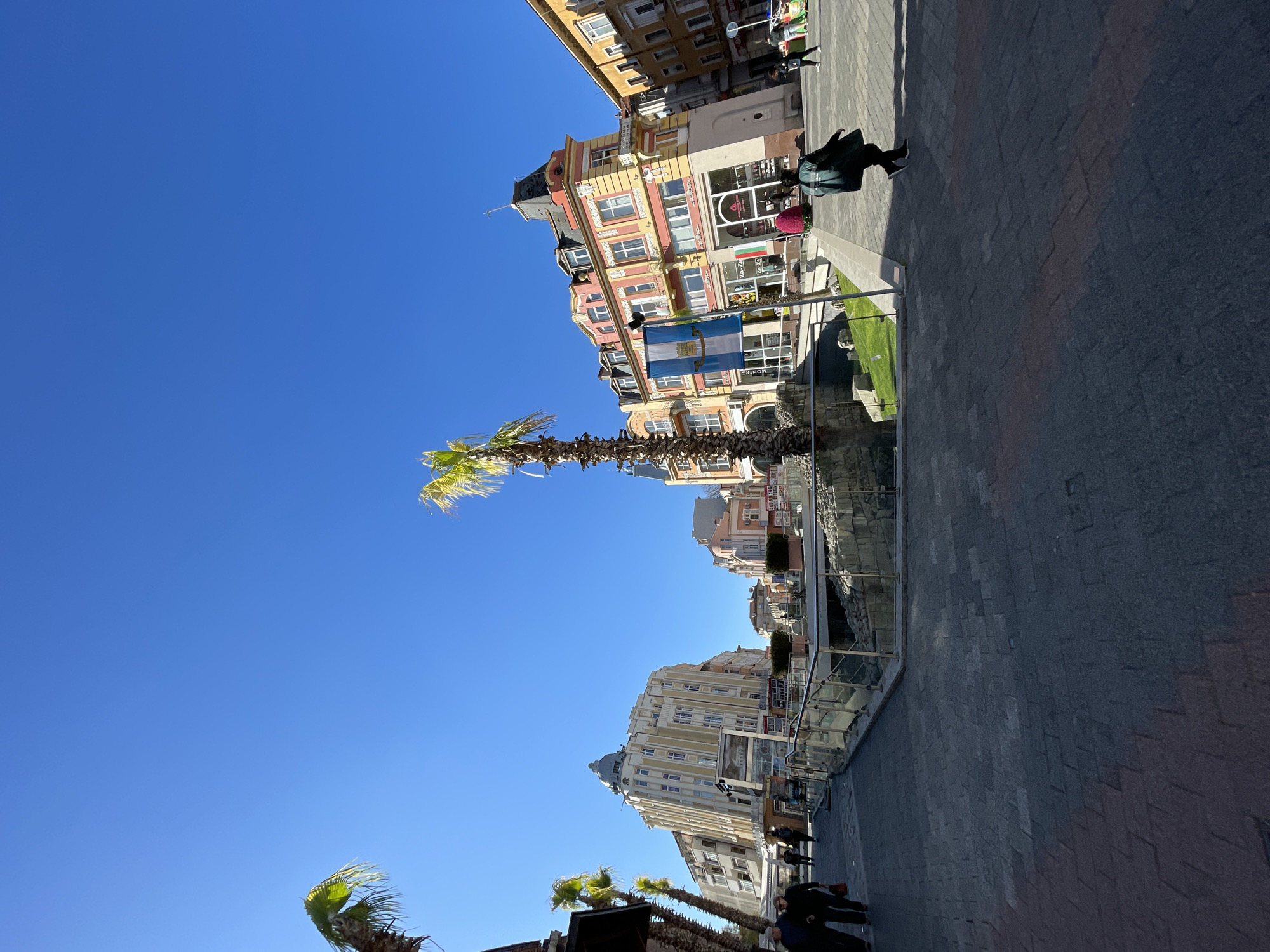 Here is a description of the image:
The image shows a street scene in Plovdiv, Bulgaria, featuring a palm tree-lined promenade. The camera angle is tilted upwards towards the clear blue sky. On either side of the promenade are buildings with architectural details, including multiple windows, ornate facades, and varying heights. Pedestrians can be seen walking along the promenade and near the buildings. A glass barrier surrounds some excavated stone ruins. The surface of the walkway is paved with patterned stones. A flag with the coat of arms of the Plovdiv region is displayed prominently on a building.