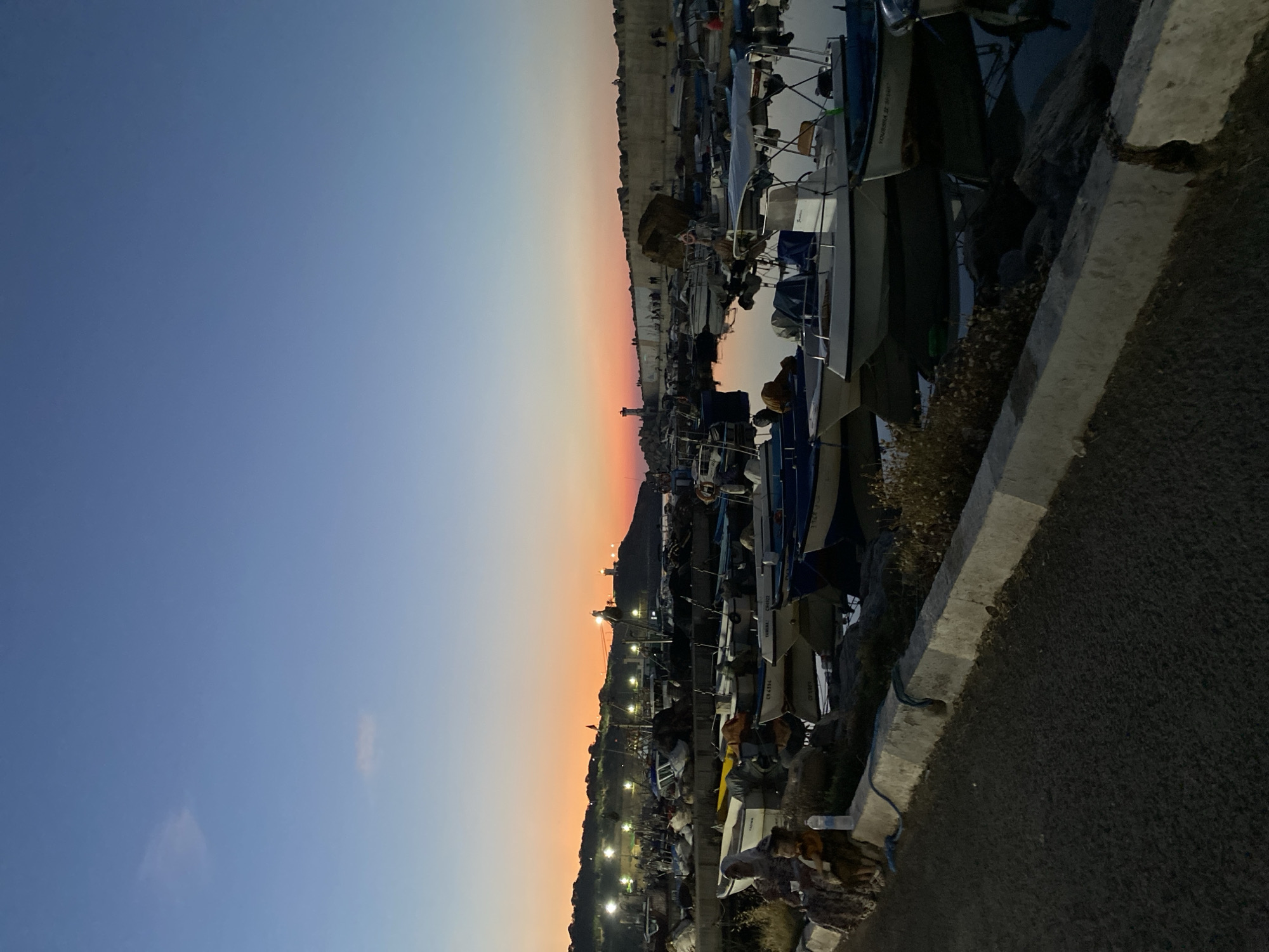 The image depicts a harbor scene at dusk in Tipaza, Algeria. The sky is a gradient of deep blue to orange and pink, indicating sunset. Numerous fishing boats are docked in the calm water, their reflections visible. In the background, a hill is illuminated by artificial lights, with a lighthouse visible on the crest. The foreground shows a paved walkway and a concrete barrier, with two people sitting on the left. The overall atmosphere is tranquil and serene.