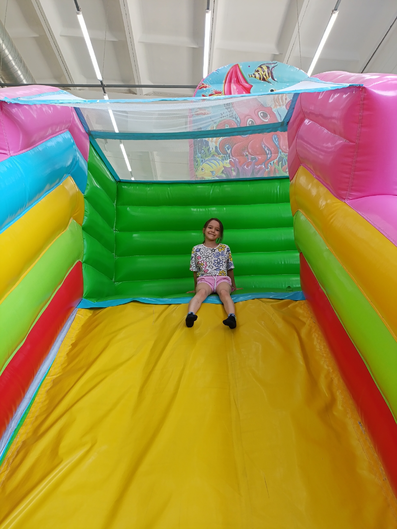 A young girl sits at the top of a brightly colored, inflatable slide. The slide's colors are red, yellow, green, blue, and pink. The slide is located inside, under bright fluorescent lights that are suspended from a white ceiling. The slide has a marine theme, with images of fish and sea creatures on the top. The girl wears a short-sleeved shirt with a floral print and pink shorts. Her feet are covered in dark socks. The location is possibly in Chaikovsky, Russia, though no specifics are definitively evident in the image itself.