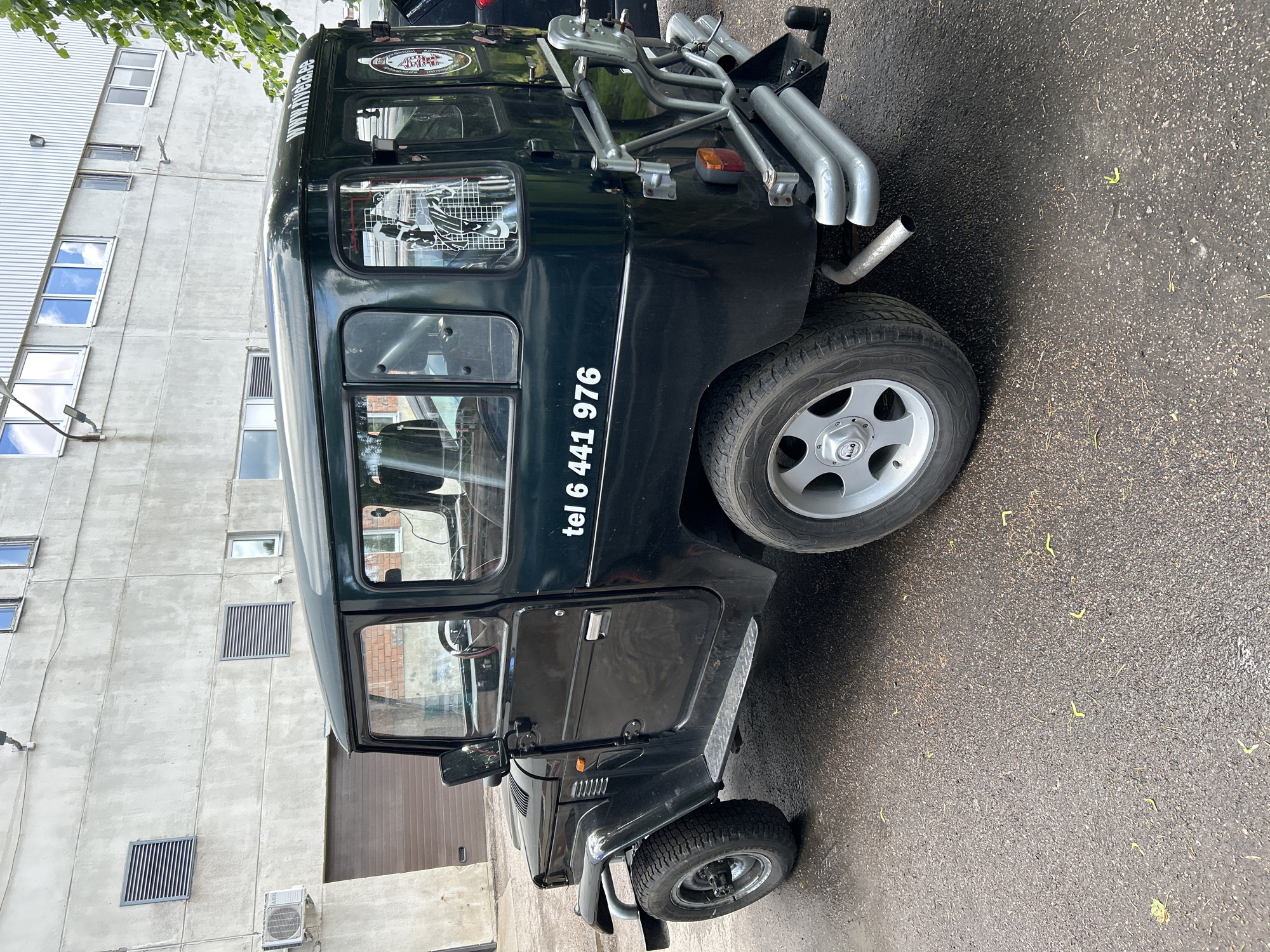 A black four-door Toyota Land Cruiser is parked on a wet asphalt surface in front of a light gray concrete industrial building. The building has a corrugated metal section on the upper part and several windows, some with horizontal louvers. An air conditioning unit is mounted to the lower left of the building wall. The car has a roof rack, a spare tire mount on the rear, and silver-colored bumpers and trim. White text on the rear door of the car reads "tel 6 441 976". A logo is visible on the rear quarter panel. Yellowish plant debris is scattered on the ground around the vehicle. The scene appears to be an outdoor area adjacent to a commercial or industrial property.