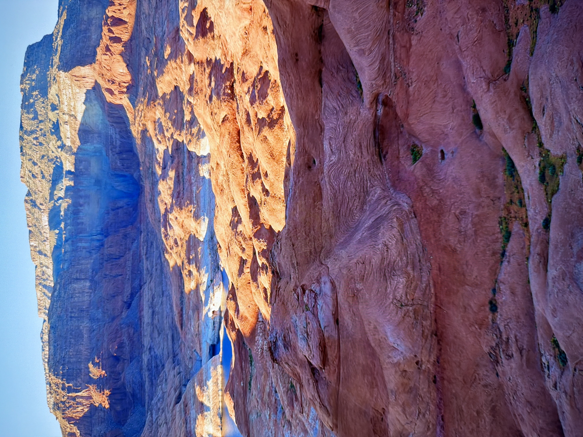 The image shows a vast, arid landscape dominated by red rock formations and a body of water. The foreground consists of undulating, eroded sandstone in shades of red and purple, with some sparse green vegetation. In the middle ground, there are more rounded rock formations and a section of bright blue water, likely a lake or reservoir, reflecting the sky. The background features towering cliffs and mesas with stratified layers, illuminated by the warm light of a setting or rising sun, casting long shadows. The sky is a clear, pale blue. There are no people, animals, or man-made structures visible in the image, and it does not depict any location in the city of Kyiv, Ukraine. The scene is natural and geological in nature.