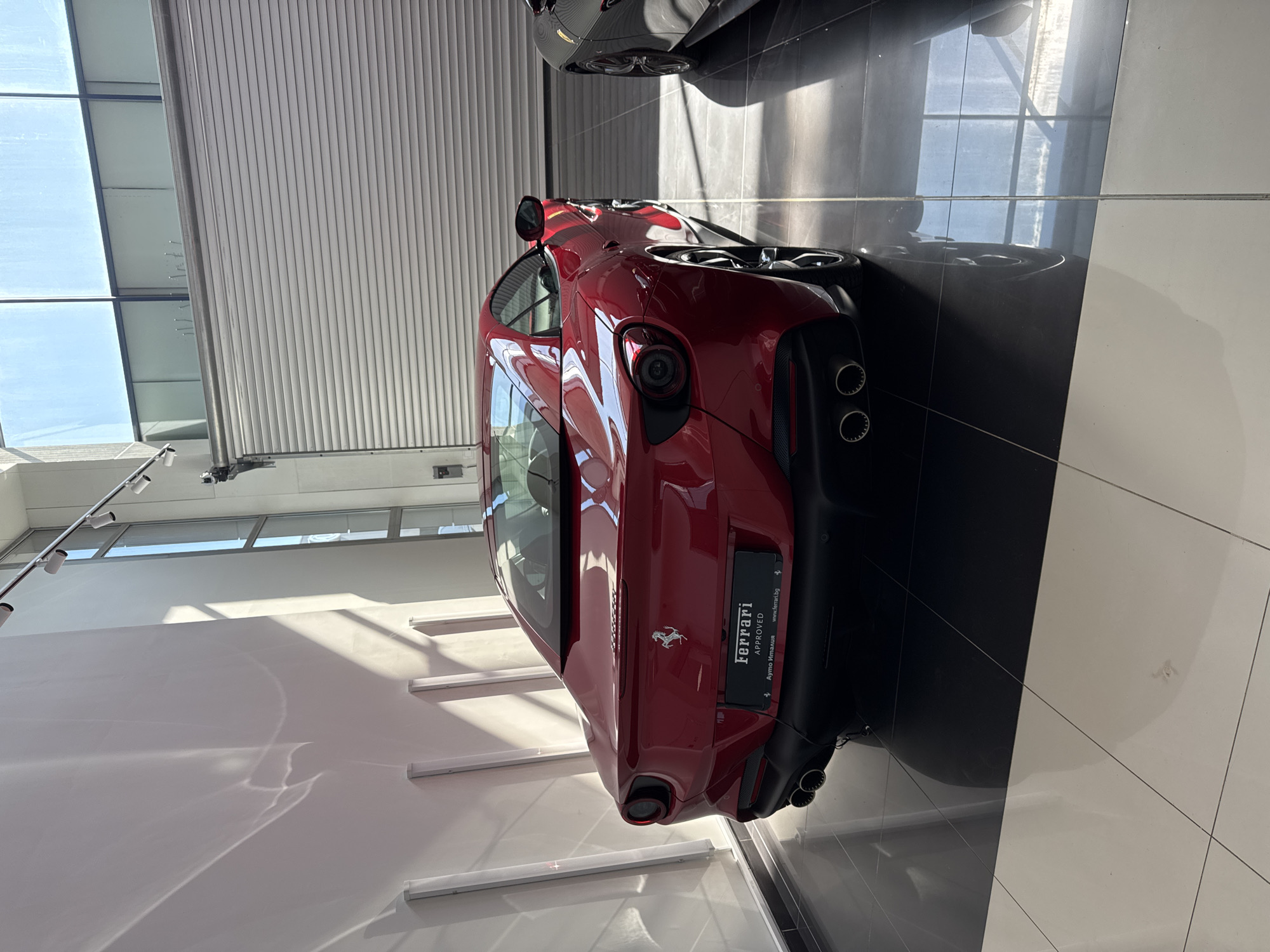 The image captures the rear of a red Ferrari parked inside a dealership showroom. The car's model is visible, bearing the "Ferrari APPROVED" badge and a Bulgarian website address ("www.ferrari.bg"), indicating the location in Bulgaria. The showroom environment is characterized by a modern design, with a white wall reflecting sunlight and a glossy black and white tiled floor. The lighting is bright, likely due to large windows, creating a stark contrast between light and shadow within the scene. Another dark vehicle is partially visible in the background.