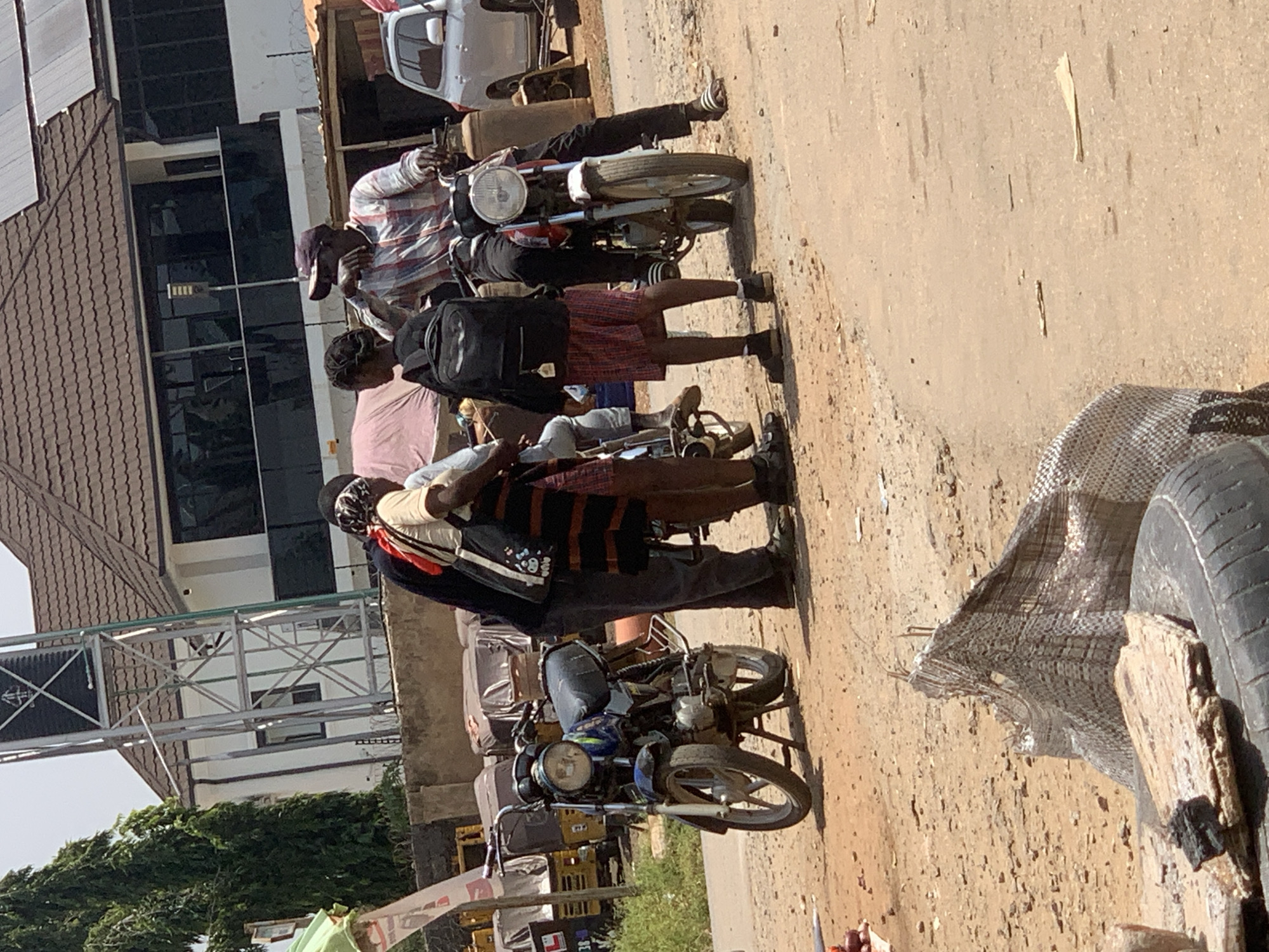 The image depicts a street scene in Abuja, Nigeria, with a focus on a group of people interacting near motorcycles. In the foreground, two motorcycles are visible. The one on the left is blue and parked on its kickstand. The one on the right has a rider wearing a plaid shirt and a dark baseball cap, and is also parked. Three individuals are gathered around the motorcycles, with one person standing behind the blue motorcycle, appearing to be in conversation with the rider of the other motorcycle. Another individual, a younger person wearing a black backpack and red and white checkered shorts, stands between the two motorcycles. The background shows a building with a tiled roof and dark windows, a tall metal structure, and several parked vehicles, including what appear to be auto-rickshaws. The ground is dusty and unpaved, typical of a street in an urban area. The overall impression is that of a casual gathering or stop along a road.