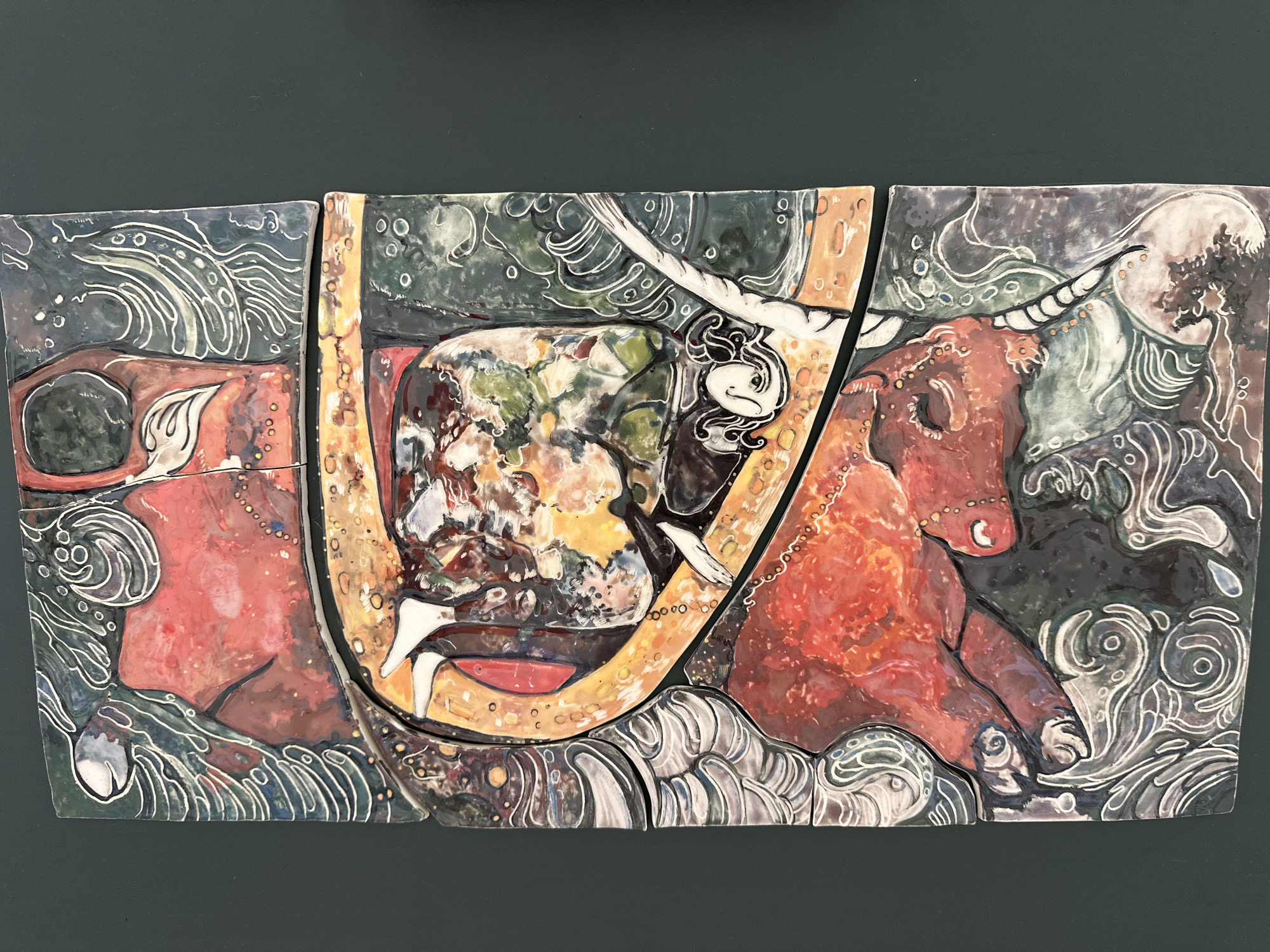 This ceramic tile artwork, located in Minsk, Belarus, depicts a vibrant and symbolic scene. The composition is formed from multiple irregular tiles, creating visible seams. On the right, a large, recumbent bull-like creature is prominent, rendered in rich red and reddish-brown tones, with visible horns and an eye. Its form suggests strength and earthy presence.
To the left, another large, abstract form in similar red hues appears, possibly representing a stylized human or animal figure, or part of a larger anatomical structure. In the center, an irregular, swirling mass of vibrant colors—greens, yellows, browns, and whites—dominates, suggestive of a world, a complex organ, or an energetic core. Emerging from the top of this central mass is a stylized white figure, resembling a serene female face or spirit, with flowing hair. Another smaller, delicate white figure, possibly a bird or a humanoid, is situated towards the bottom-center.
Throughout the artwork, white, flowing lines and patterns are intricately drawn against a dark green and blue background, which may represent water, sky, or an ethereal plane. These lines give a sense of movement, energy, or perhaps stylized waves or clouds. The overall style is mythological and folk-art inspired, utilizing a rich palette and glossy ceramic glazes. No text is visible in the image.