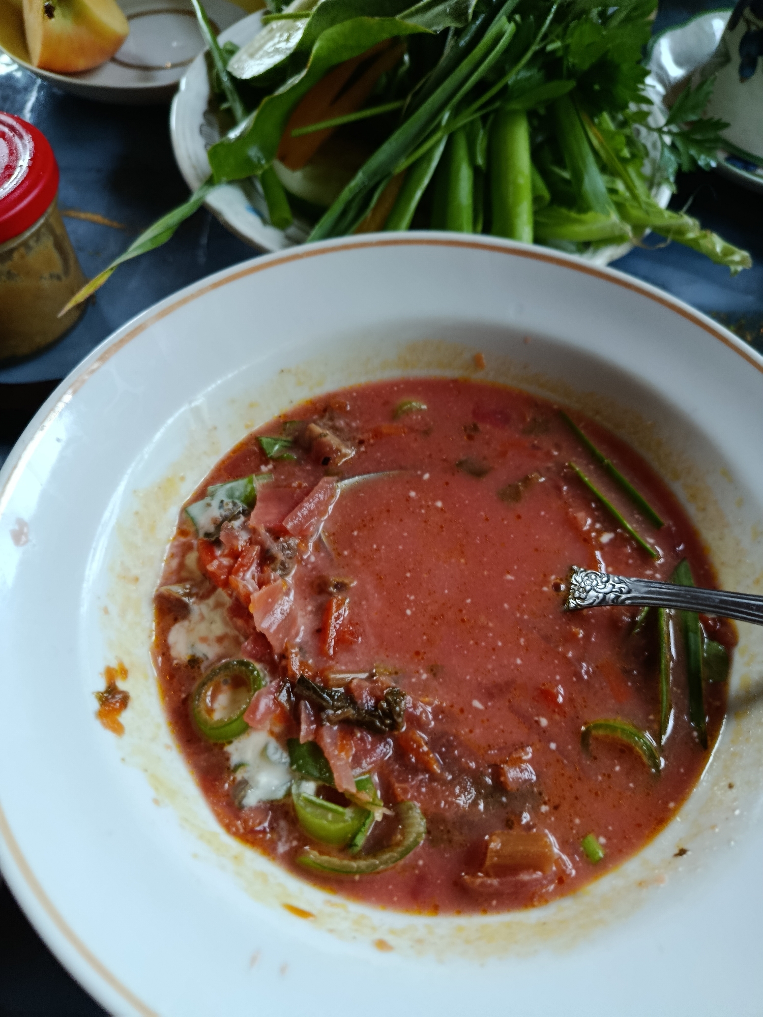A close-up, overhead shot captures a bowl of borscht, a traditional Eastern European soup, on a dark surface. The soup is a vibrant red, with visible chunks of vegetables such as beets, cabbage, and possibly tomatoes. Slices of green onion are scattered throughout the soup, and a dollop of white cream or sour cream is present. A silver spoon rests in the bowl, its ornate handle suggesting it is part of a set. To the left of the bowl, a small glass jar with a red lid is partially visible, containing a yellowish-brown substance, possibly a condiment or spread. In the background, a white plate holds a pile of fresh green vegetables, including long stalks of what appears to be scallions or leeks, and leafy greens. A slice of yellow fruit, possibly an apple or persimmon, is also on this plate. The overall setting suggests a home-cooked meal, likely in Верея, Russia, given the context. The lighting is natural, casting soft shadows and highlighting the textures of the food.