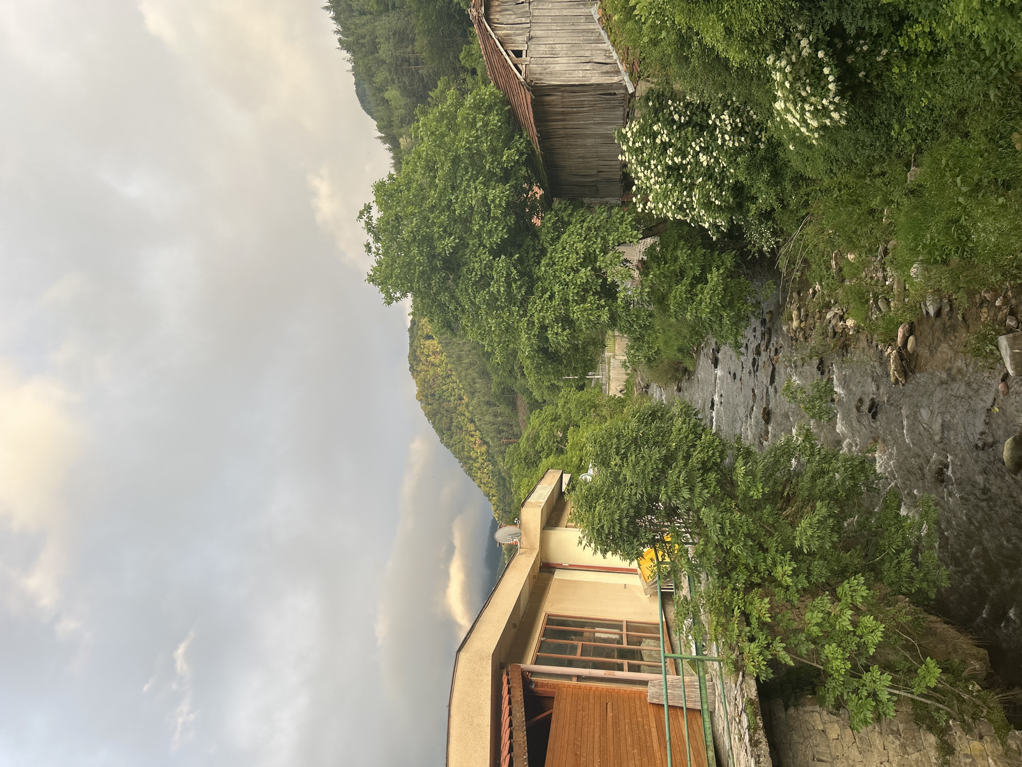 The image captures a serene landscape featuring a river, lush vegetation, and two buildings under an overcast sky. The perspective is slightly elevated, looking down towards a clear, stony river that flows from the background towards the foreground.
On the far bank of the river, to the right, an old, dilapidated two-story wooden building stands out. It has weathered wooden walls and a red-tiled roof, part of which appears damaged. Large, dense green trees and bushes with small white flowers surround this structure, blending it into the verdant hillside behind, which is densely forested with dark green trees.
In the foreground, on the left bank, a more modern, two-story building is visible. It features a light-colored facade, a flat roof with a satellite dish, and prominent wooden-framed windows. The lower section of this building has vertical wooden paneling, and a green metal railing borders its edge near the river. A stone wall forms the base of this modern building.
The sky is heavily overcast with thick, grey and white clouds, suggesting a cloudy day with diffused light. There are no people visible, and no specific activities are taking place, contributing to a calm and quiet atmosphere. The scene combines natural beauty with a blend of old and newer architectural styles, hinting at a rural or mountainous setting. No visible text is present.