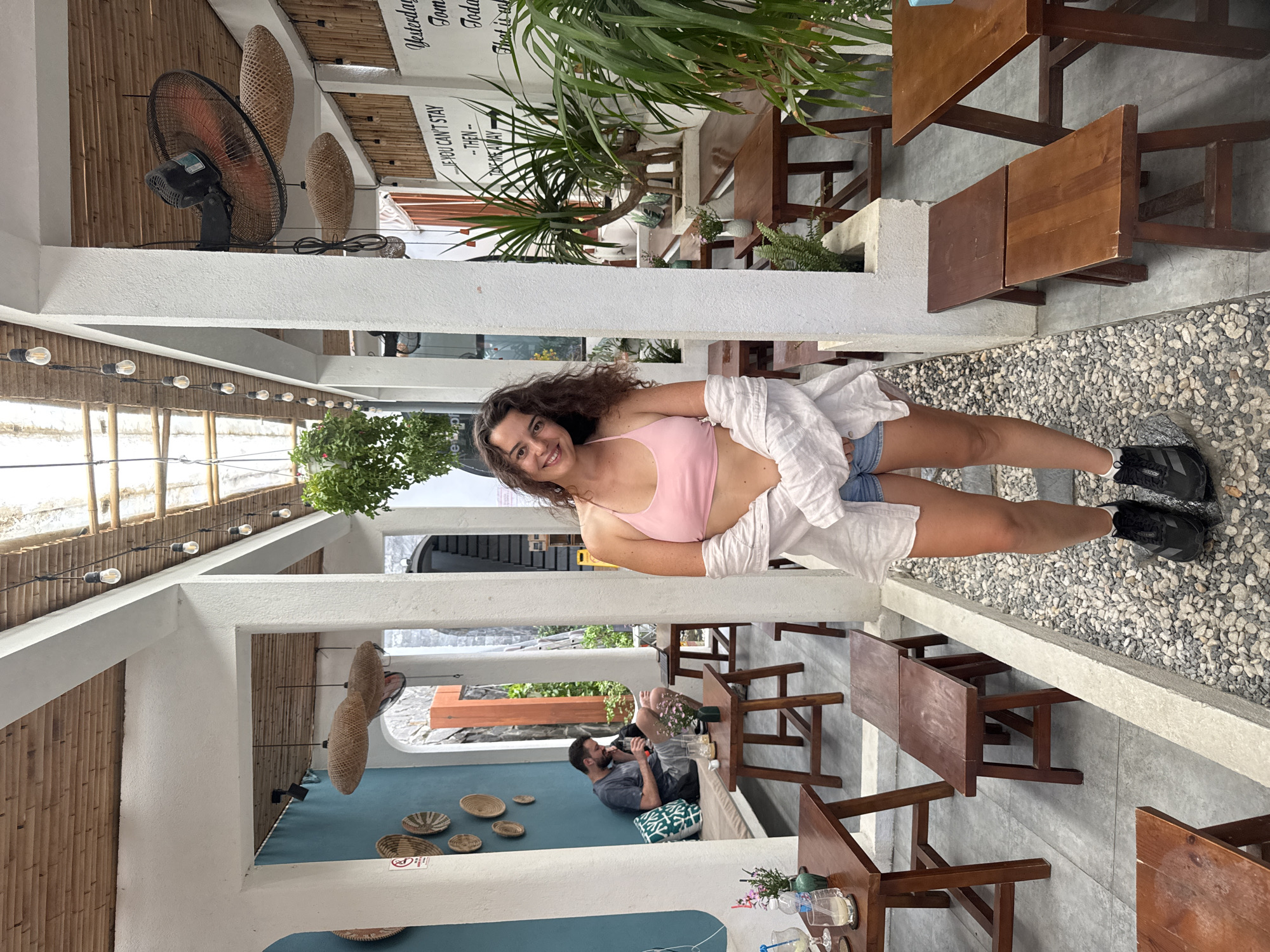 In a cafe in Hoi An, Vietnam, a woman stands in a pathway between tables and benches. She has long, curly brown hair and is wearing a pink bikini top, denim shorts, and a white unbuttoned shirt. She is also wearing black athletic shoes. The cafe has a rustic interior with a bamboo ceiling, white pillars, and wooden furniture. The pathway is covered in gravel. In the background, there is another person sitting on a bench, a fan mounted on the ceiling, and plants. There is also writing on the wall in English.