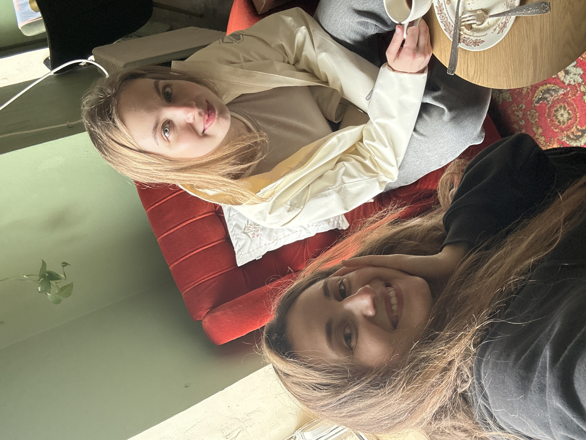Two young women are seated in an indoor setting. One, with blonde hair and fair skin, is wearing a white jacket and grey pants. The other, with light brown hair, is wearing a dark top. They are sitting on a red couch near a small round wooden table with a plate, utensils, and a coffee cup on it. The setting appears to be a cafe or home with a green wall and a red patterned rug. The city of Kryvyi Rih, Ukraine, is not discernible from the image.