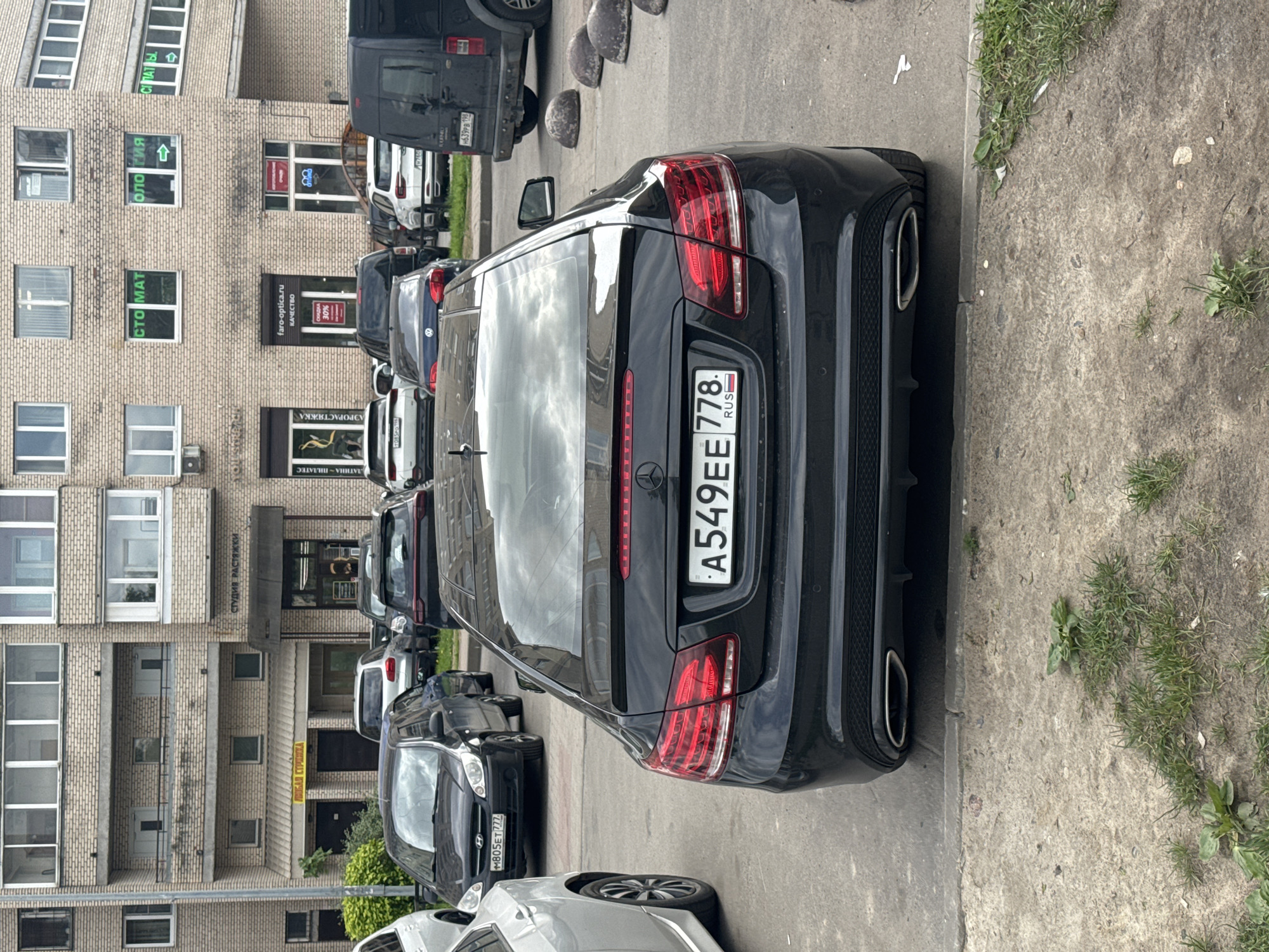 The image depicts an urban street scene in Saint Petersburg, Russia, under an overcast sky during daylight hours. Several cars are parked along a curb, adjacent to a multi-story brick building.
The main subject is a black Mercedes-Benz sedan, parked in the foreground and viewed from its rear-left side. Its Russian license plate, `A549EE 778 RUS`, is clearly visible. Behind it, a black Hyundai SUV is parked, displaying the license plate `M805ET 777`. Further down the street, a white van has the license plate `H639PB 198`. The cars are stationary, with no human activity immediately apparent.
The brick building houses various businesses on its ground floor, identified by Cyrillic signage. Notable texts include "СТУДИЯ РАСТЯЖКИ" (Stretching Studio), "ЛАТИНА ПИЛАТЕС" (Latina Pilates), and "АЭРОРАСТЯЖКА" (Aerial Stretching). Other signs read "CTO MAT" (likely part of "Стоматология" - Dentistry) and another partial sign displaying "ология" (suggesting a medical or dental practice) next to an arrow. A prominent sign for an optical store, "faro-optica.ru", advertises "КАЧЕСТВО СКИДКА 30%" (QUALITY DISCOUNT 30%). Another sign at the top reads "С ПАТЫ." with an arrow. The street features a concrete curb, patchy grass, and rounded concrete bollards used for traffic management. The dark cars reflect the cloudy sky.