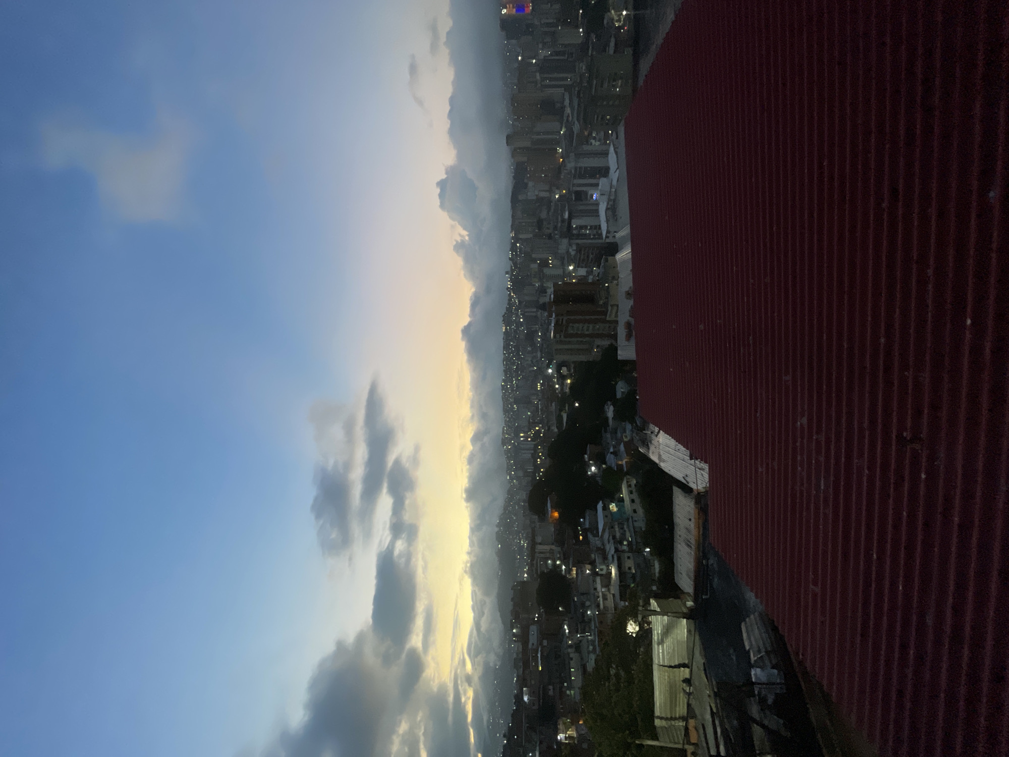 Here is a description of the image:
The image presents an aerial view of Caracas, Venezuela, at dusk or dawn. The sky transitions from a light blue with scattered clouds to a hazy yellow-orange near the horizon, where the urban landscape begins. The cityscape is densely populated with buildings of varying heights, illuminated by artificial lights. Some structures are tall enough to resemble skyscrapers. The urban area extends into the distance, seemingly nestled within mountainous terrain. The foreground shows corrugated metal roofing.