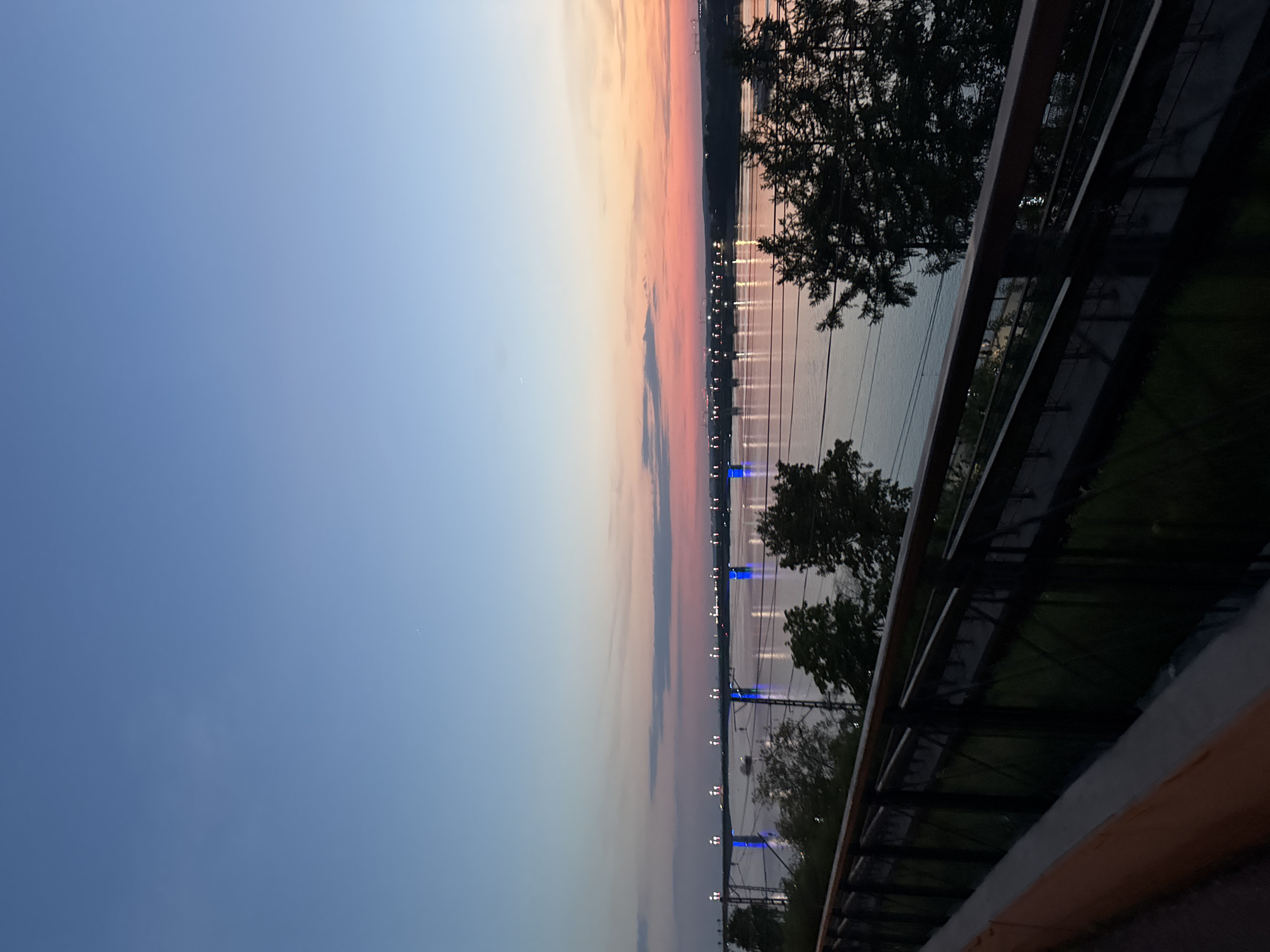 The image shows a sunset over the Kama River in Perm, Russia. The sky transitions from blue to pink and orange, and the river reflects the lights of the city. A bridge spans the river, with its supports illuminated in blue. A fence and trees are visible in the foreground.