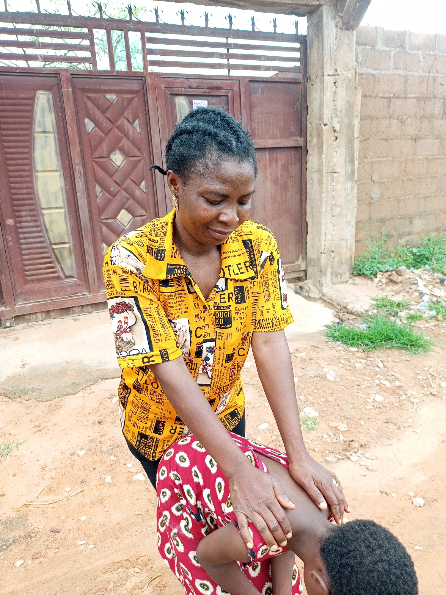 In Tempure, Nigeria, a woman with braided hair and wearing a yellow patterned shirt kneels to tend to a child who is bent over in front of her. The woman's hands are gently placed on the child's back. The setting appears to be outdoors, with a dirt ground, a brick wall, and a large metal gate with wooden accents in the background. Some green vegetation and scattered debris are visible to the right of the scene.