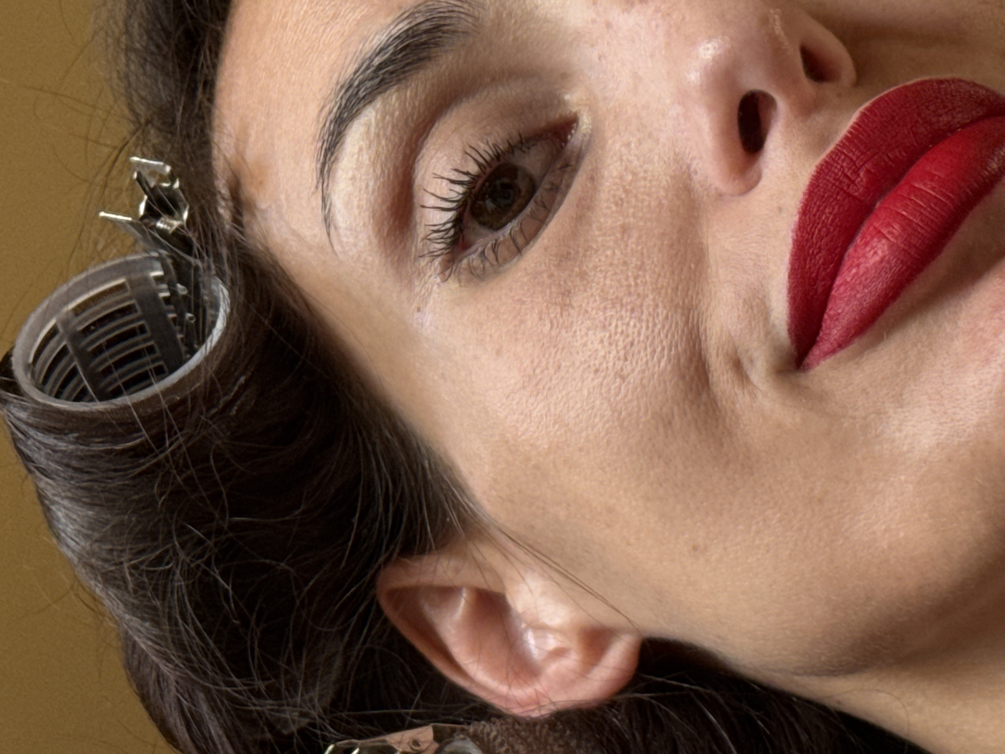 The image is a close-up, diagonal portrait of a woman's face, focused on her right eye, nose, and mouth, along with her hair styled in rollers. Her head is angled upwards and towards the right side of the frame, occupying most of the visual space.
The woman has dark, well-defined eyebrows and a prominent brown eye, which is open and looking slightly to the right. Her eyelashes appear to be enhanced with mascara. Her skin displays natural texture, including visible pores and subtle lines, particularly around the eye and cheek, along with a few faint freckles. Her lips are painted with a vibrant, matte red lipstick, slightly parted in a subtle smile or thoughtful expression, creating a gentle dimple or crease near the corner of her mouth. Part of her nose is visible on the right side of the frame.
Her dark brown hair is in the process of being styled, with several plastic hair rollers secured by metal clips visible on the left side of her head, framing her ear. Some loose strands of hair are also apparent.
The background is a plain, warm-toned color, appearing as a light brown or ochre wall, suggesting an indoor setting such as a home or a salon. The activity depicted is hair preparation or styling, possibly for an event. The woman's expression is calm and neutral, with a hint of a soft, understated smile. There are no explicit location cues within the image itself, though the provided context is Pleven, Bulgaria. No text is visible in the media file.