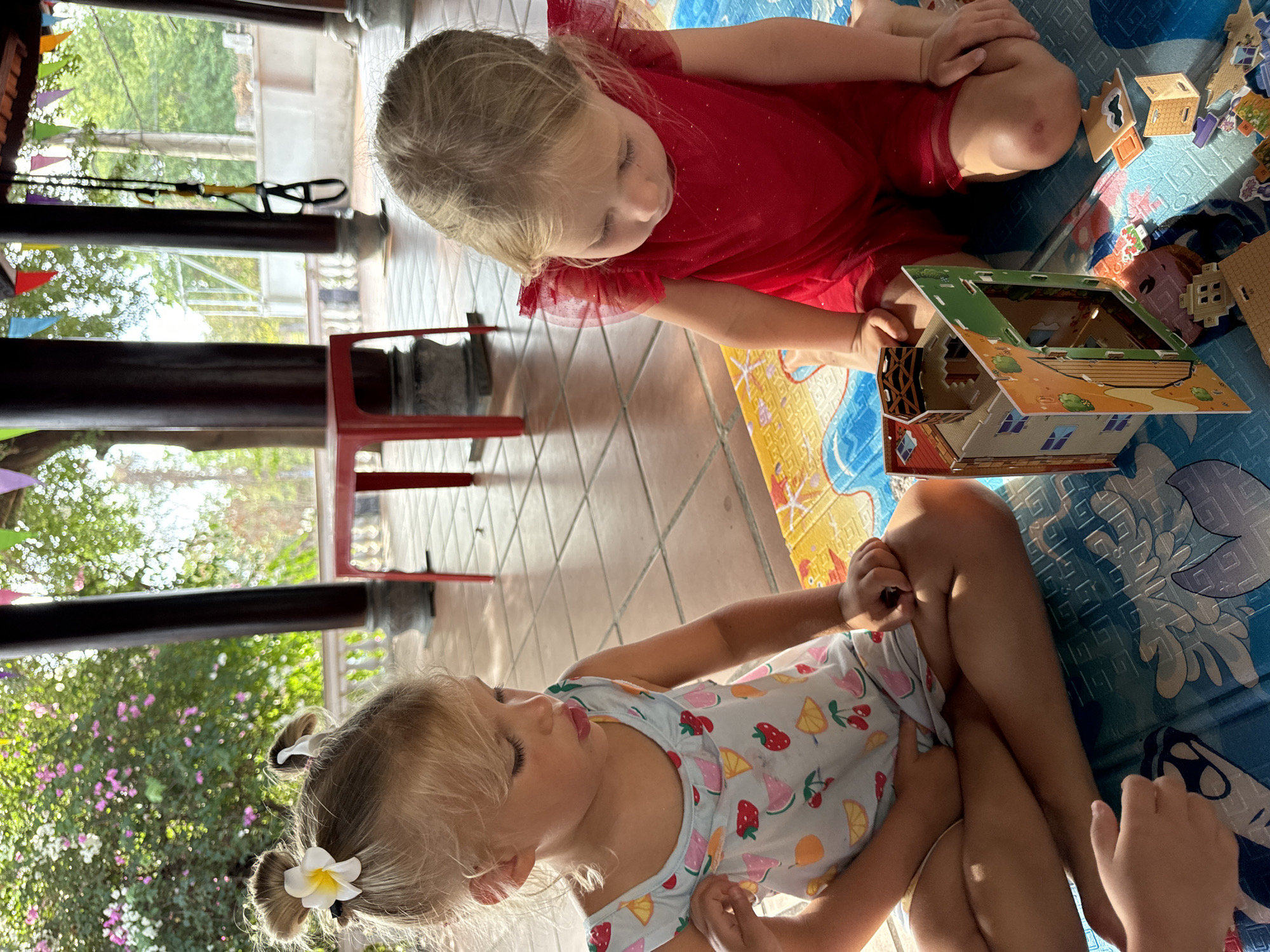Two young girls are seated indoors on a blue patterned mat, assembling a cardboard model house. The girl closest to the camera wears a pale blue dress printed with fruit designs and has a white flower in her hair, styled in pigtails. She sits cross-legged, her attention focused on the project. The other girl wears a red dress with tulle detailing and is leaning towards the house model. The house is partly assembled, showing cardboard sections of walls, windows, and a green roof. Additional cardboard pieces are scattered on the mat. The floor is tiled. The scene appears to be within a building in Mũi Né, Vietnam, evidenced by the exterior view of foliage and colorful triangular flags visible through a window. Dark beams and a red stool are also visible in the background.