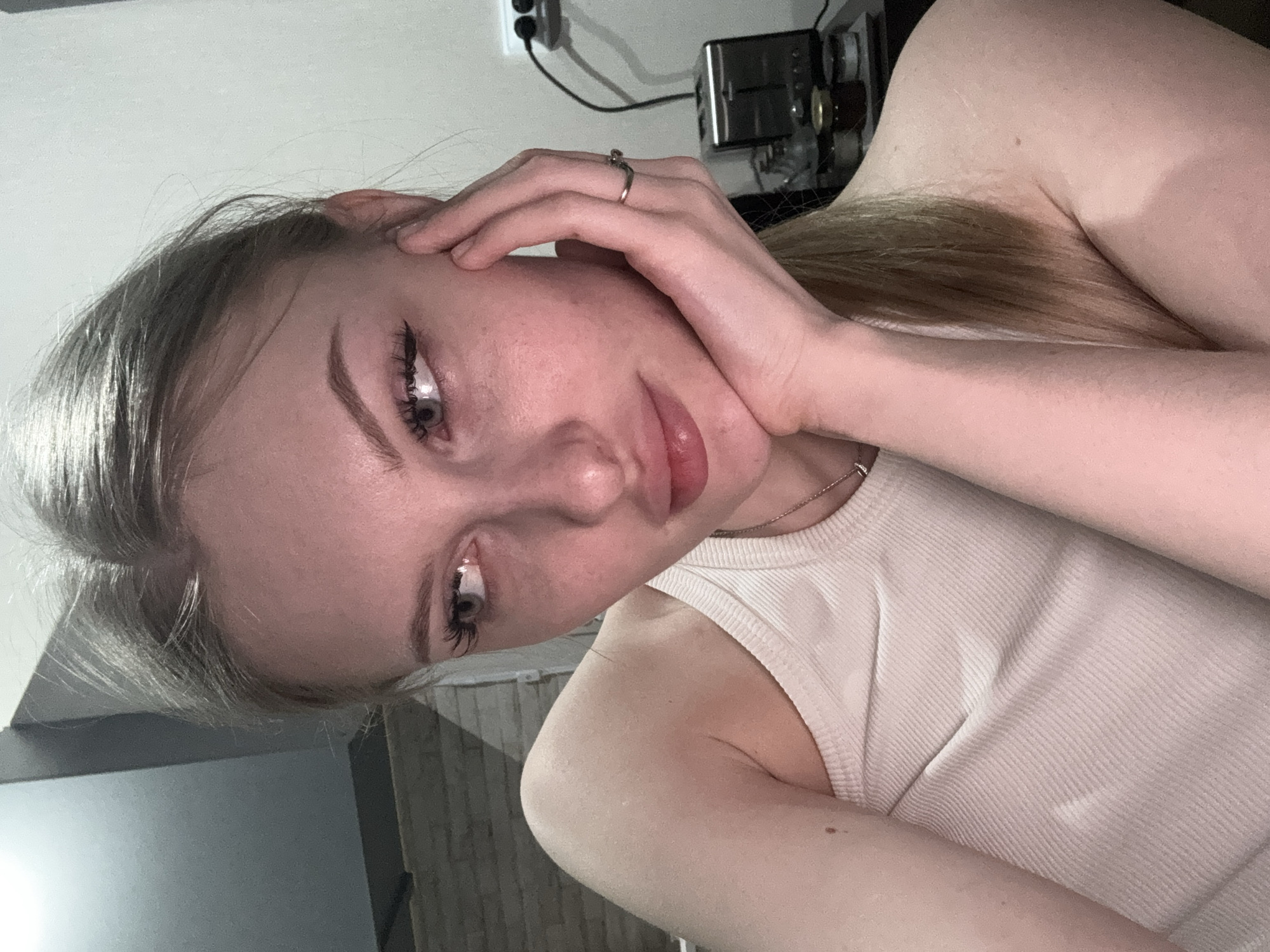 A young woman with fair skin and light brown hair is lying down, with her head tilted towards the viewer. She is wearing a white tank top and a ring on her left hand. Her hand is positioned near her face. The background shows a white wall with an electrical outlet and a small appliance. The location cannot be determined from the image.
