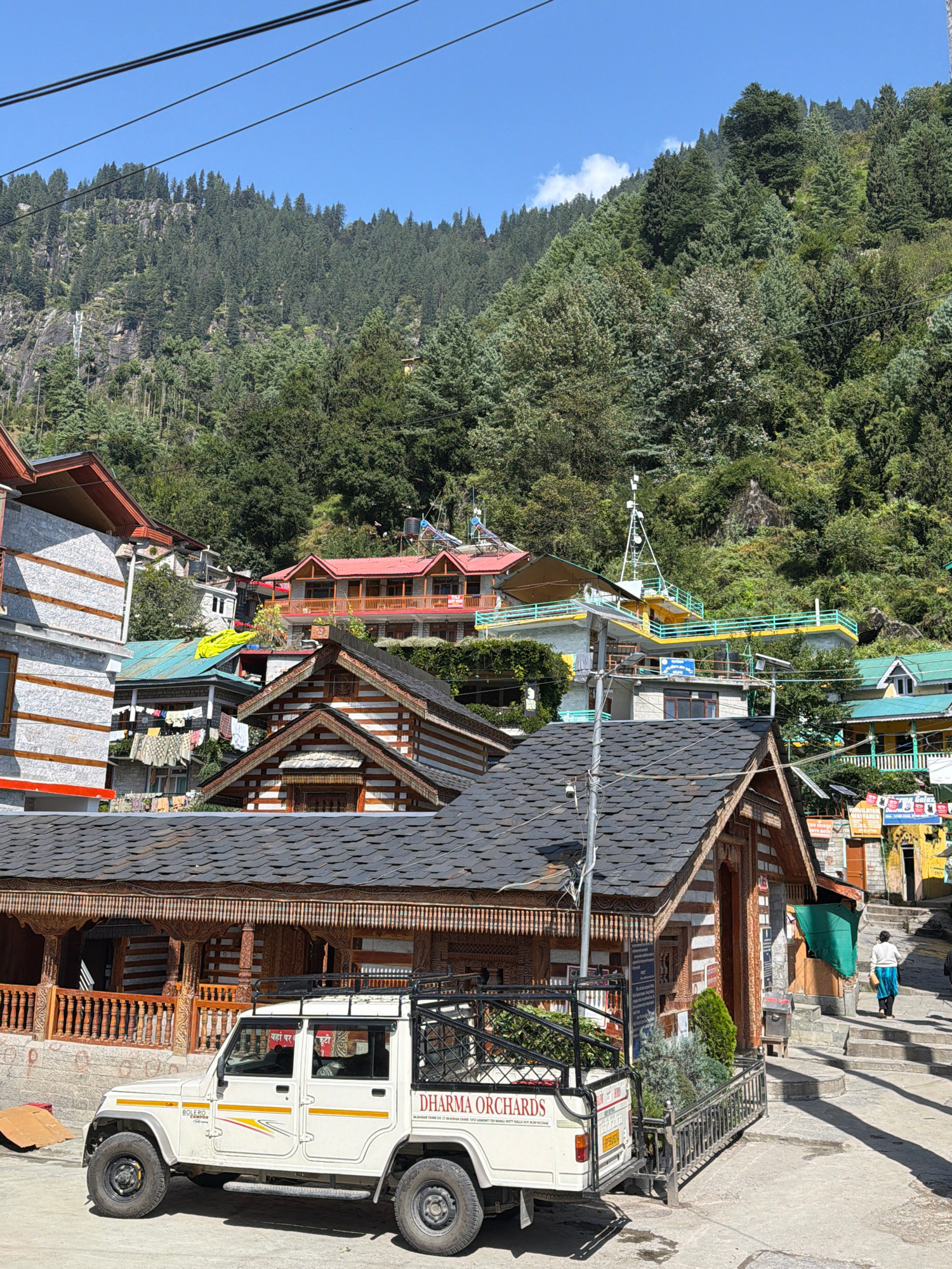 In the town of Vashisht, India, a white Mahindra Bolero pickup truck is parked on a paved street in front of a traditional wooden building with a dark, sloped roof. The truck has "DHARMA ORCHARDS" written on its side in red lettering. Behind the truck and building, multiple houses with red and green roofs are visible, built on a steep hillside covered in dense pine trees. A narrow, paved pathway with stairs leads upwards from the street, where a person in a white top and blue skirt is walking away from the camera. The sky is clear and blue.