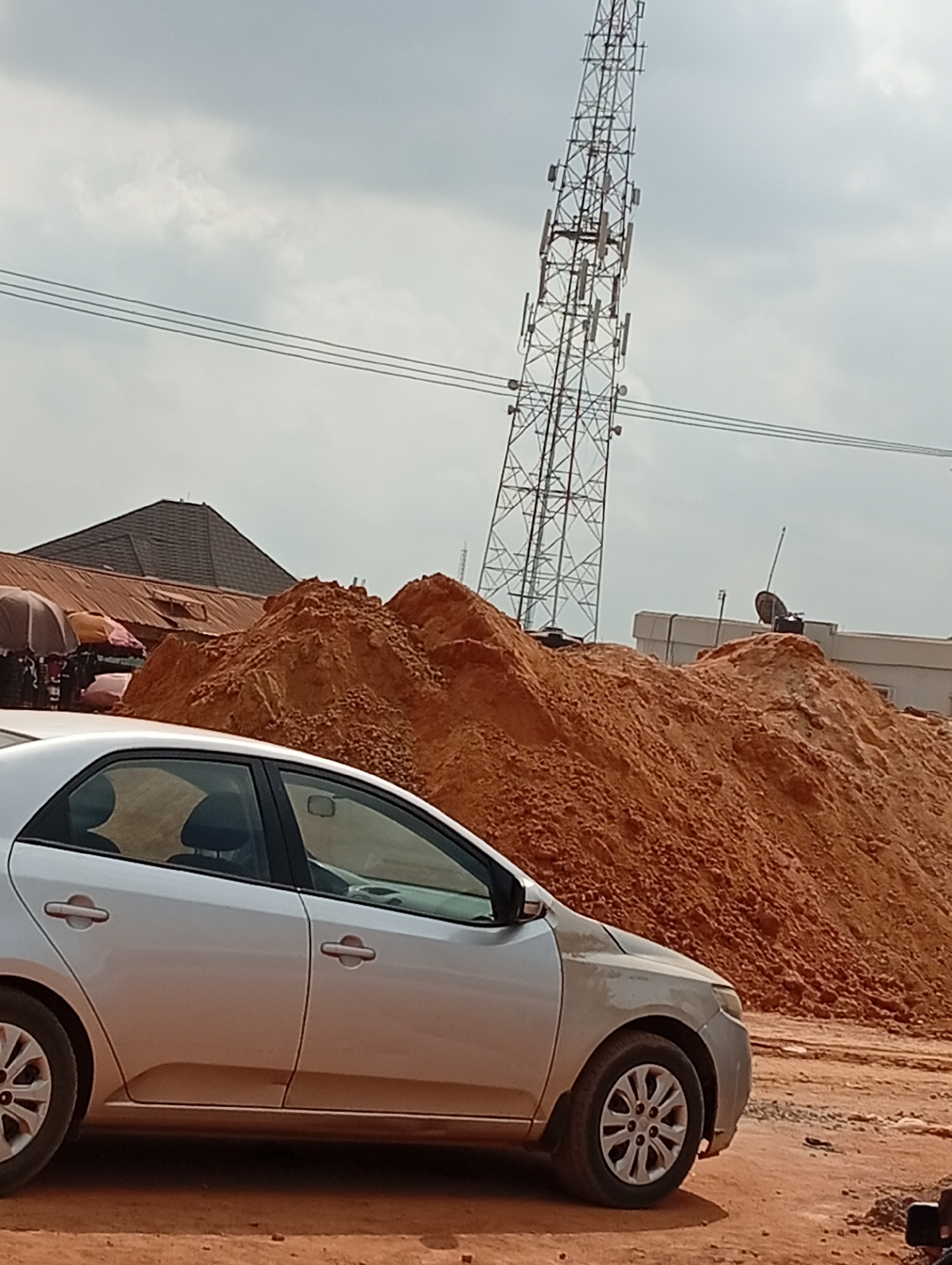 The image shows a silver sedan parked on a reddish-brown dirt lot in Ado, Nigeria. A large pile of reddish-brown earth rises behind the car. In the background, a tall communications tower is visible, as well as parts of buildings, including some with rusty roofs, and tents. The sky is overcast. No people are clearly visible in the image.