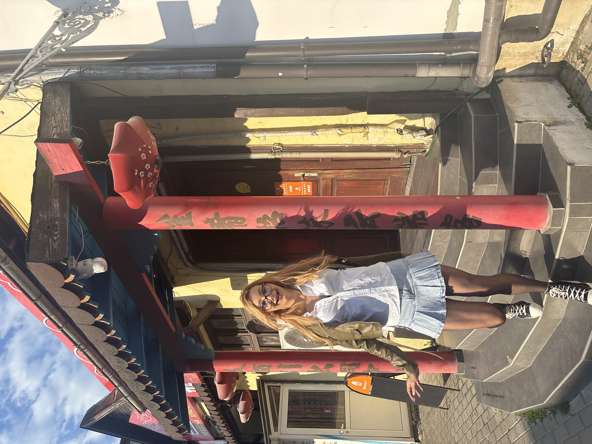 A blonde woman wearing glasses, a white blouse, a denim skirt, a green jacket, black stockings, and black and white sneakers is posing under a red Chinese archway in Varna, Bulgaria. The archway is decorated with Chinese characters and red lantern-like ornaments. Behind the archway is a weathered yellow building with a brown wooden door and exposed pipes. A security camera is visible on the side of the arch. To the side of the woman is a stairway. The sky with clouds is visible to the side.