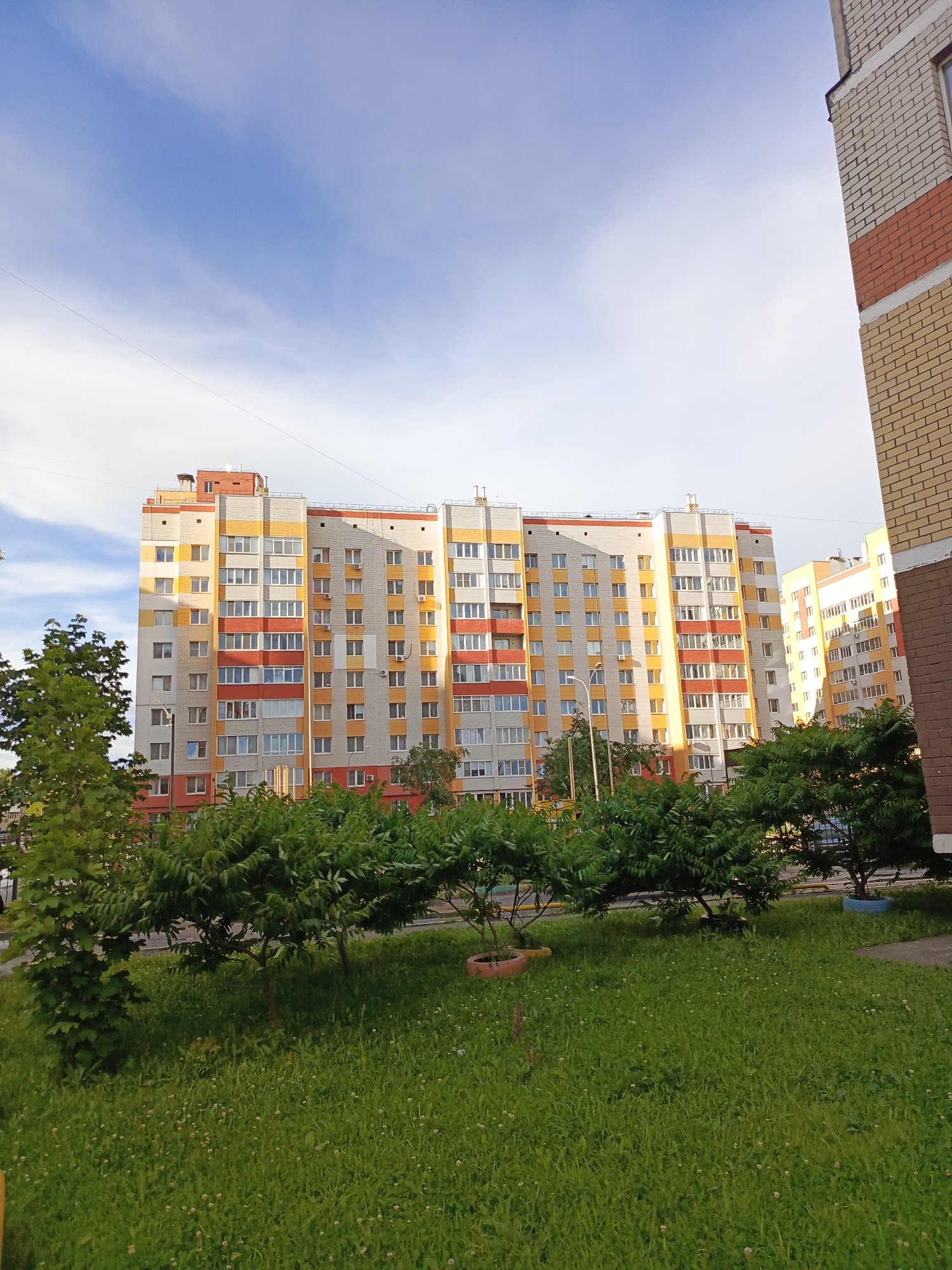 This media file depicts an outdoor scene in Bryansk, Russia, likely during the daytime. The primary subject is a modern, multi-story apartment building with a colorful facade, featuring sections of yellow, orange, and red brickwork interspersed with white panels and numerous windows. To the right, another apartment building, partially visible, shares a similar architectural style.
In the foreground, a well-maintained grassy area is dotted with small white flowers. Several lush green trees and bushes are planted in front of the apartment buildings, partially obscuring their lower sections. One tree in the foreground is more sparsely leaved, while others are dense and leafy. There are also two planters visible in the grass, one orange and one blue, adding a touch of color to the landscape. A lamppost stands near the apartment building.
The sky above is a bright blue with scattered white clouds, suggesting fair weather. The lighting indicates it is likely late afternoon or early evening, with the sun casting shadows. There is no discernible activity or event taking place, and no people are visible. The overall impression is a peaceful, urban residential environment. No visible text is present in the image.