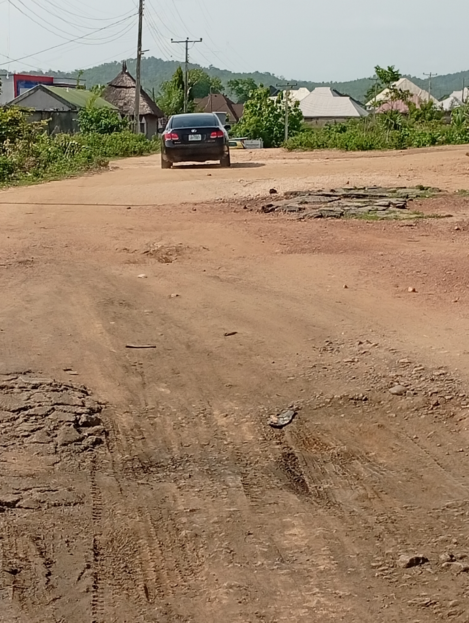 The image depicts a deteriorated dirt road in Adankolo, Nigeria. The road surface is heavily damaged with potholes and tire tracks. A black car is driving away in the distance on the road. Buildings and vegetation line the road, with a small hill visible in the background. Overhead electrical wires and poles are present.