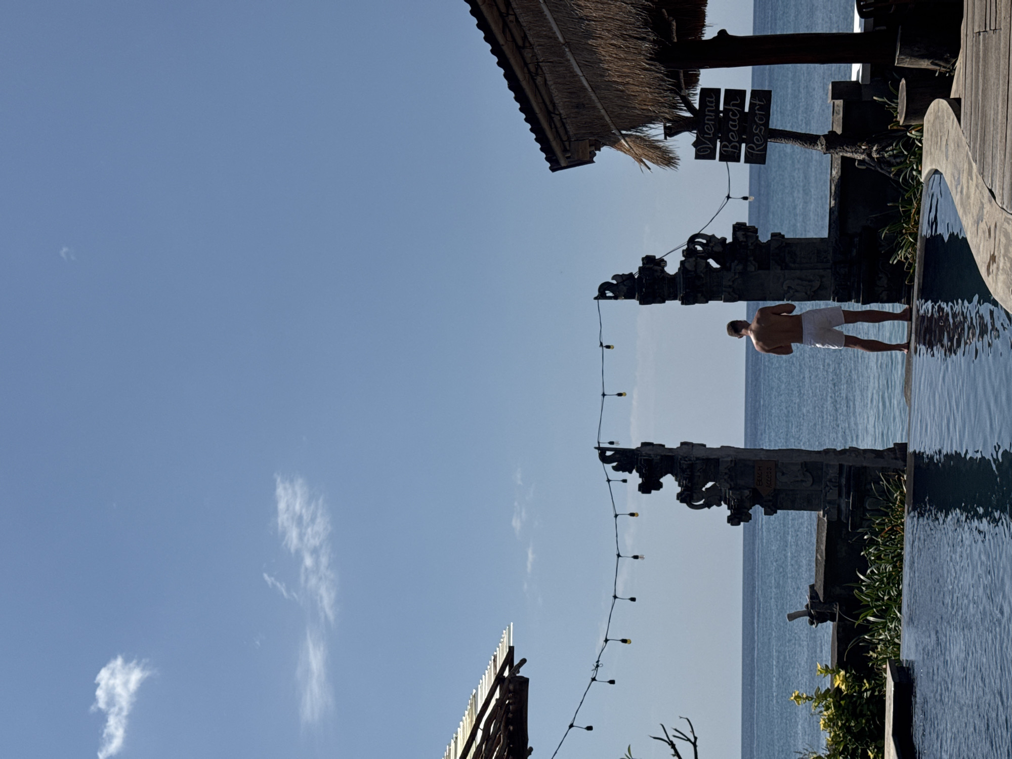 A man stands facing the ocean in Abang, Indonesia, by a resort. He is wearing white shorts and is shirtless. He is standing at the edge of an infinity pool, with the ocean visible behind him. To his right is a traditional Balinese stone gate, and beyond that, a thatched-roof structure. A sign on a post reads "Vienna Beach Resort". String lights hang between the gateposts. The sky is clear blue with a few wispy clouds.