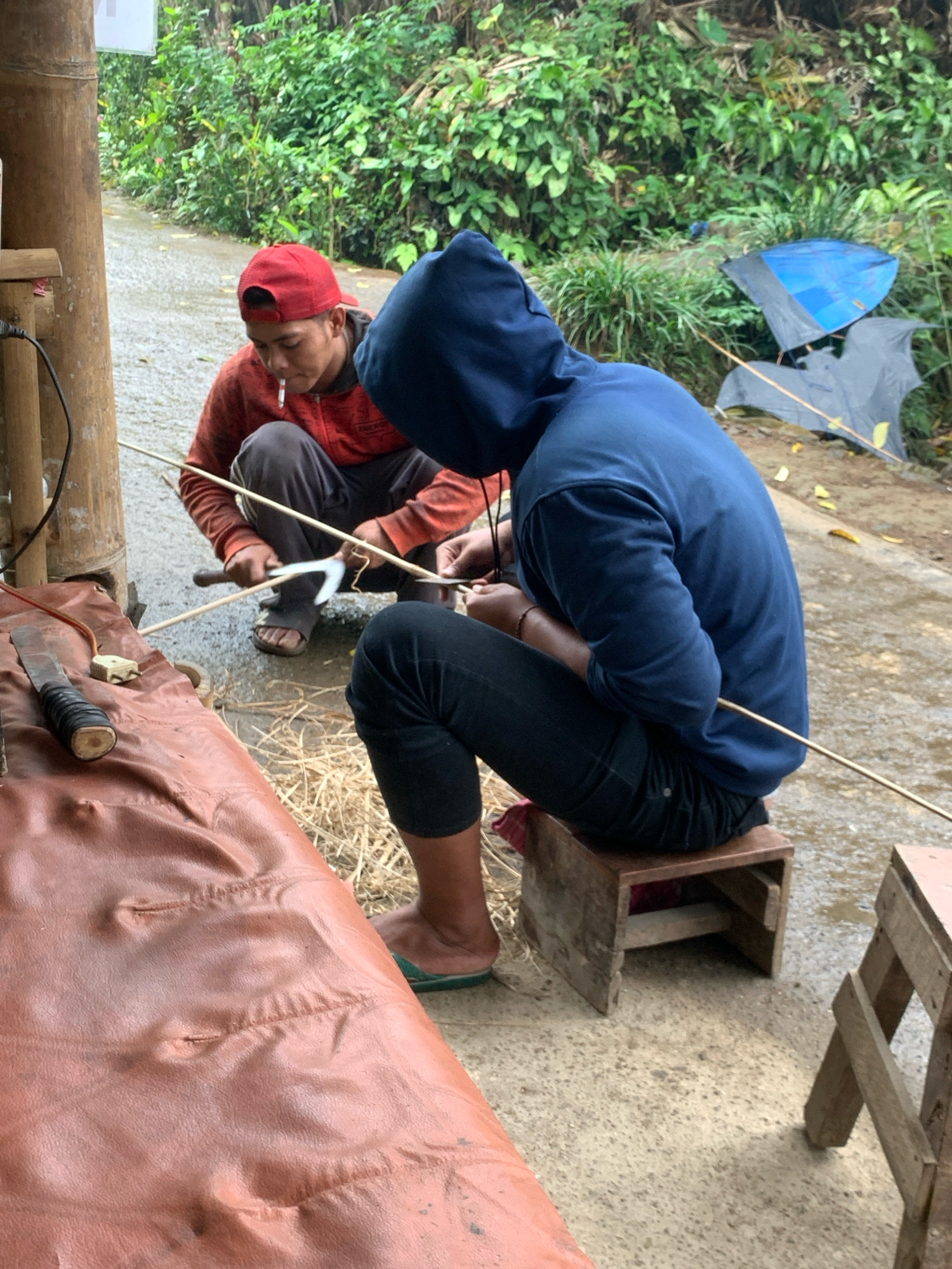 In Pronojiwo, Indonesia, two individuals are engaged in an outdoor activity near lush vegetation and a paved path. One person, wearing a red hooded jacket and a red cap, is crouched down and appears to be smoking a cigarette. They are holding a long, thin stick and a sharp tool, possibly to craft something. The second person, facing away from the camera and wearing a blue hooded jacket, is also sitting on a wooden stool and working with a similar stick. In the background, two kites, one blue and gray, are laid out on the ground. The environment suggests a rural or semi-rural setting, possibly a roadside or open area with natural surroundings.