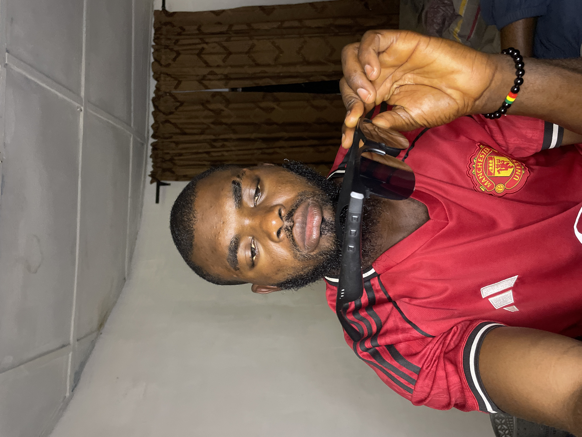 In the image, a Black man with short hair and a beard is holding a pair of sunglasses in front of his face. He is wearing a red Manchester United jersey with black stripes on the sleeves and white accents on the collar and cuffs. On his left wrist, he wears a black beaded bracelet with colored bands of red, yellow, and green. The man is indoors, in what appears to be a room with a textured ceiling and brown curtains in the background. The lighting suggests it is evening or nighttime. The overall scene is casual and personal. This image does not provide any specific visual cues or context that would allow for its placement within the city of Jos, Nigeria.