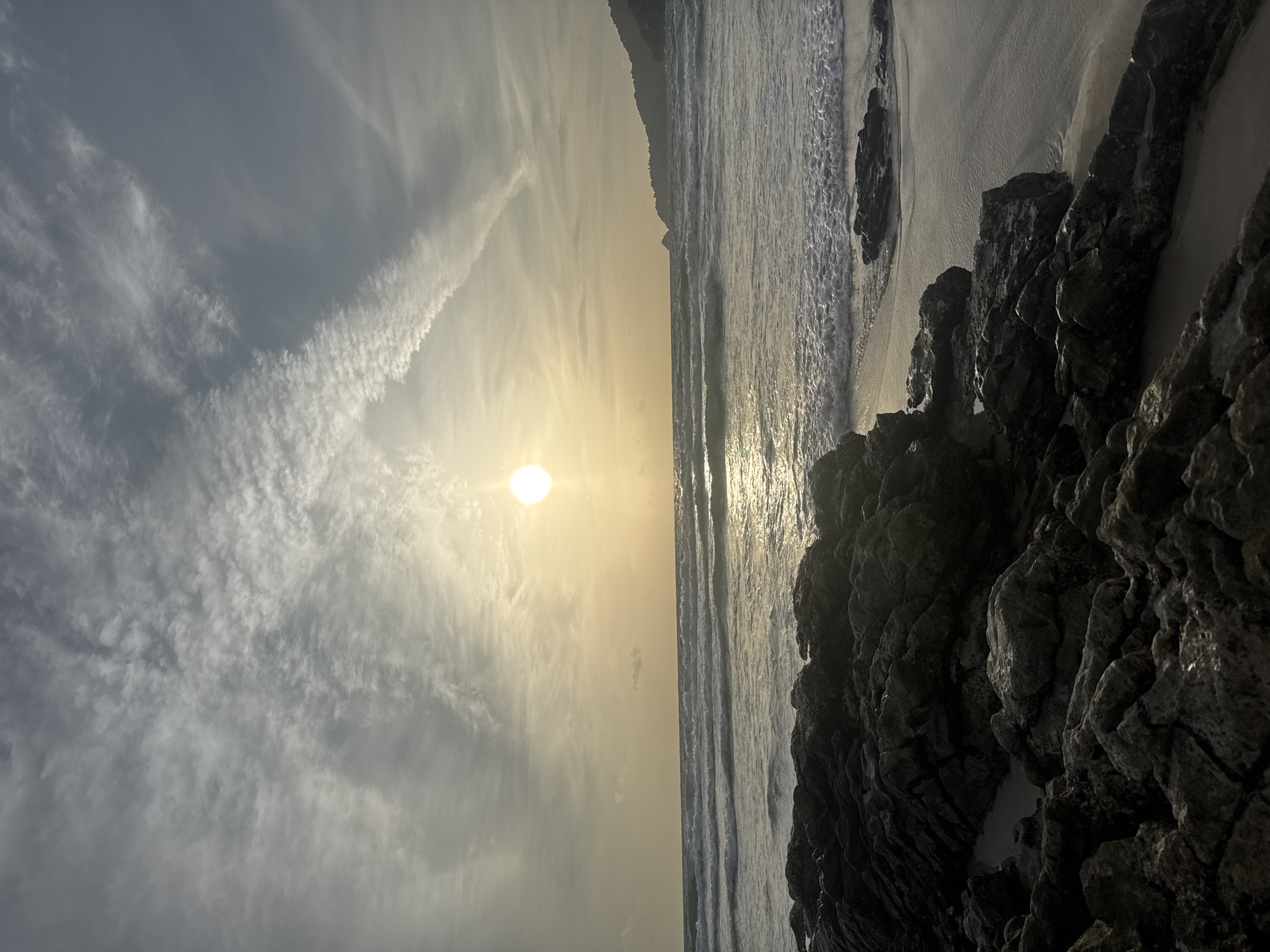 A serene beach scene in Patong, Thailand, captures the golden hour as the sun dips towards the horizon. The sky is a canvas of soft, muted grays and whites, with wispy clouds stretching across the expanse. The sun, a bright orb, casts a warm glow on the water, creating a shimmering reflection. Gentle waves roll onto the sandy shore, their white foam a stark contrast to the darker, wet sand. In the foreground, dark, jagged rocks are scattered along the coastline, adding a rugged texture to the otherwise smooth landscape. The water extends to the horizon, where a faint outline of a distant hill is visible, silhouetted against the fading light. The overall mood is peaceful and tranquil, inviting contemplation of nature's beauty.