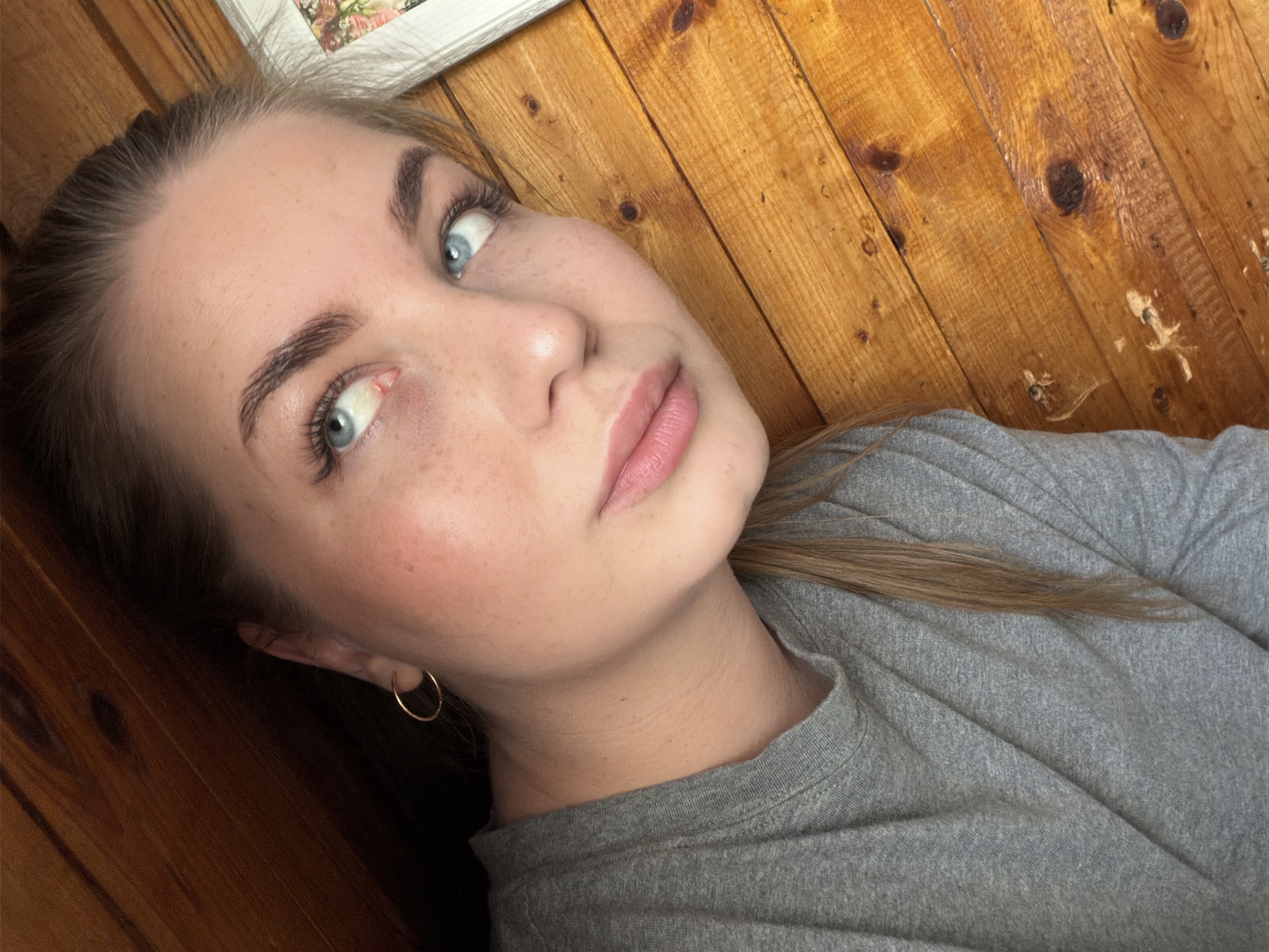 A young woman with fair skin, freckles, and light blue eyes is looking upwards at the camera. Her hair is pulled back, and she is wearing a small gold hoop earring. She is wearing a grey t-shirt. The background is a wooden wall. A picture frame is partially visible in the top left corner. The location is not discernible from the image.