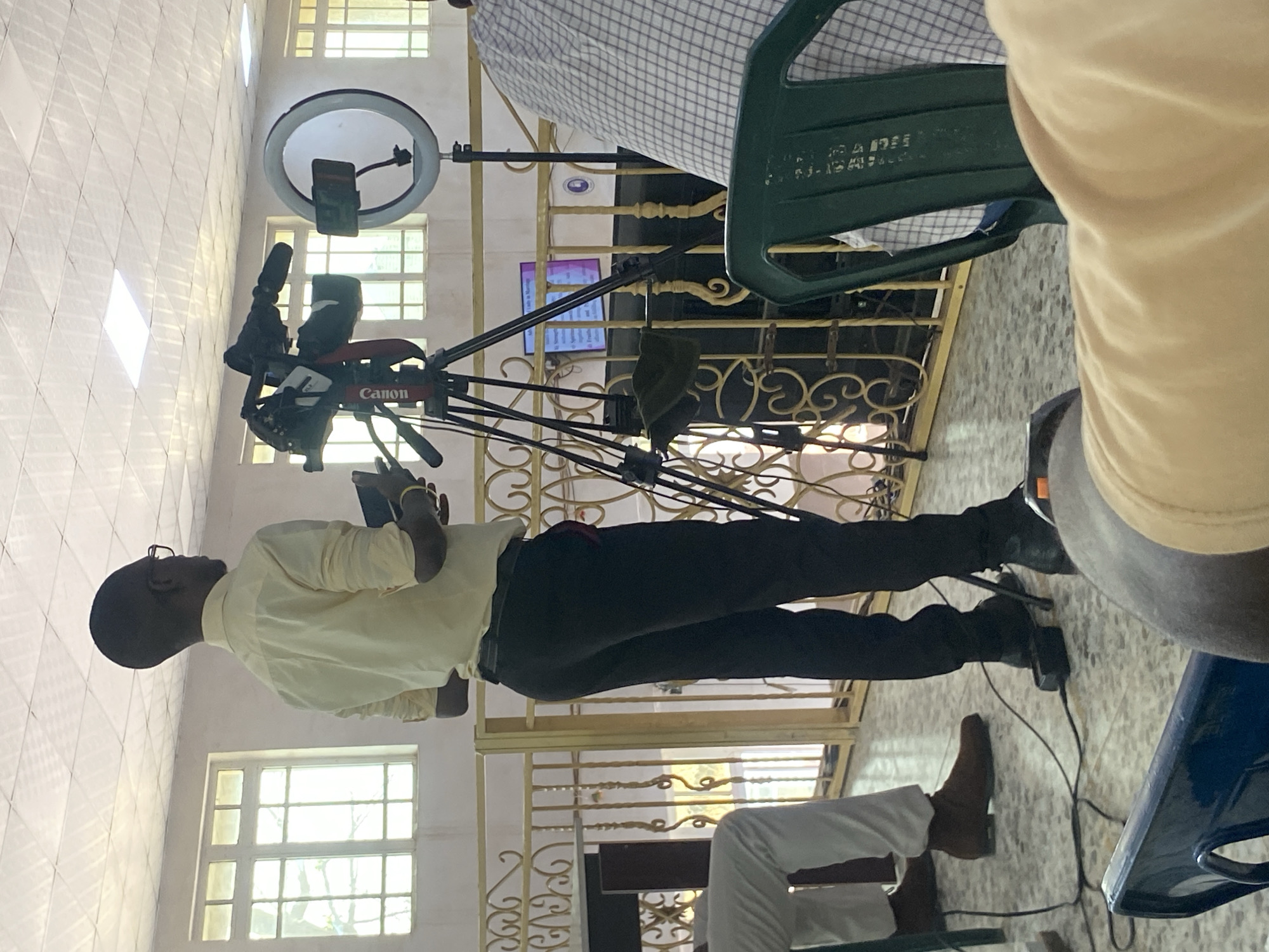 A man stands in front of a Canon video camera on a tripod, holding a phone or small tablet, seemingly operating the camera setup. The camera is positioned in front of a gold-colored, ornate railing or gate, with a screen or monitor visible behind it displaying text. A ring light is also part of the setup. The man is wearing glasses, a light-colored shirt, and dark pants with black shoes. Part of another person wearing a checkered shirt and sitting in a green chair is visible to the upper right. A second person wearing white pants and brown shoes is sitting in the lower part of the image. Windows are visible in the background with sunlight shining through. The scene is likely indoors, and judging by the architecture and details of the environment, it might be in Barden Yana, Nigeria.