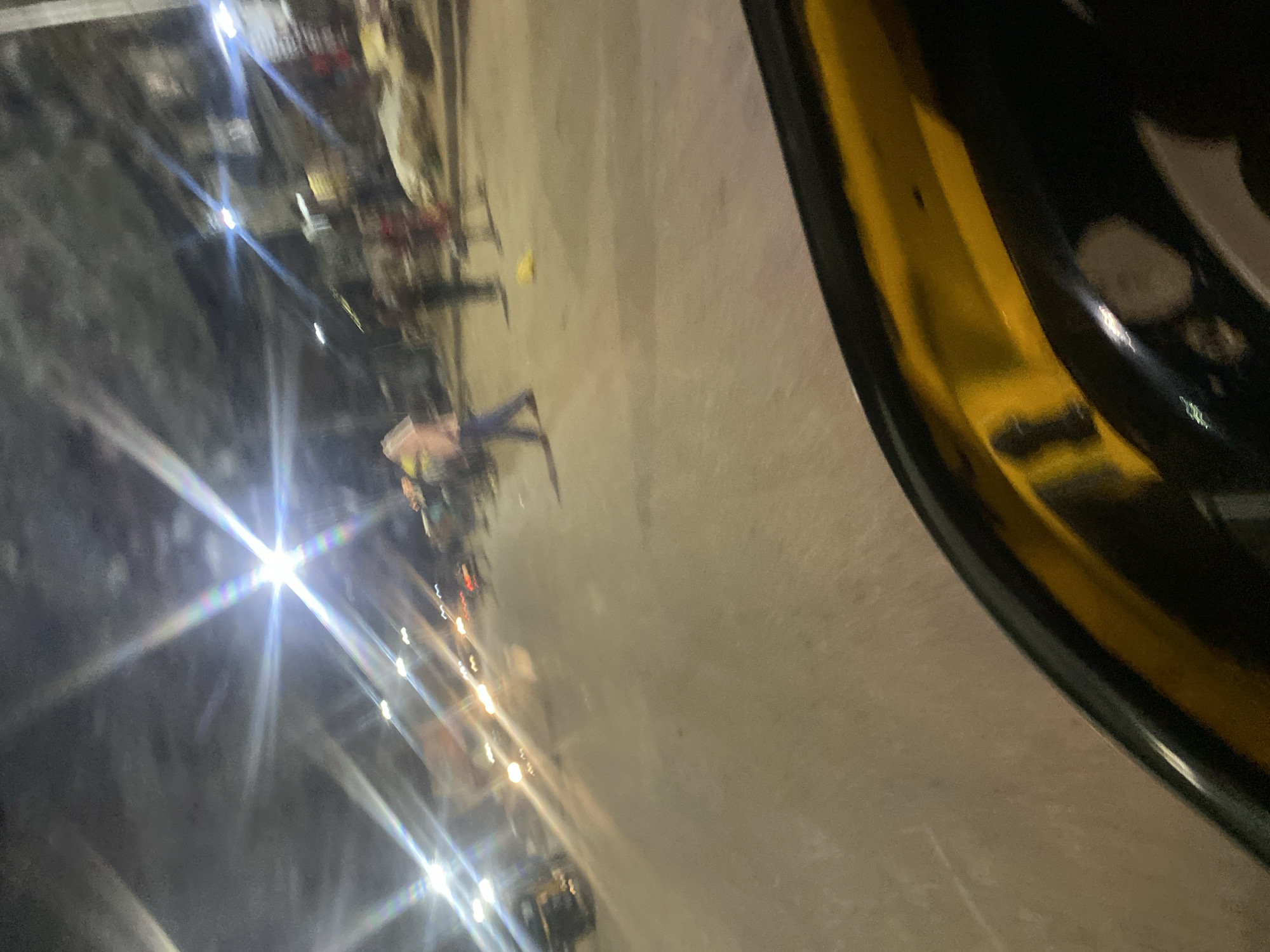 The image captures a street scene at night, possibly in Gwong, Nigeria. Bright lights from vehicles and street lamps create starburst effects due to the camera's aperture. Several blurred figures, appearing to be people, are visible walking on the street. One individual in the foreground is wearing a light-colored shirt and dark pants. Vehicles, including what appears to be a tuk-tuk or auto-rickshaw, are also present, indicated by their headlights. The road surface is visible, and there are buildings or structures lining the street, though they are indistinct due to the low light and motion blur. The foreground of the image is dominated by the dark, curved edge of a vehicle, likely the one from which the photo was taken, with a yellow accent.