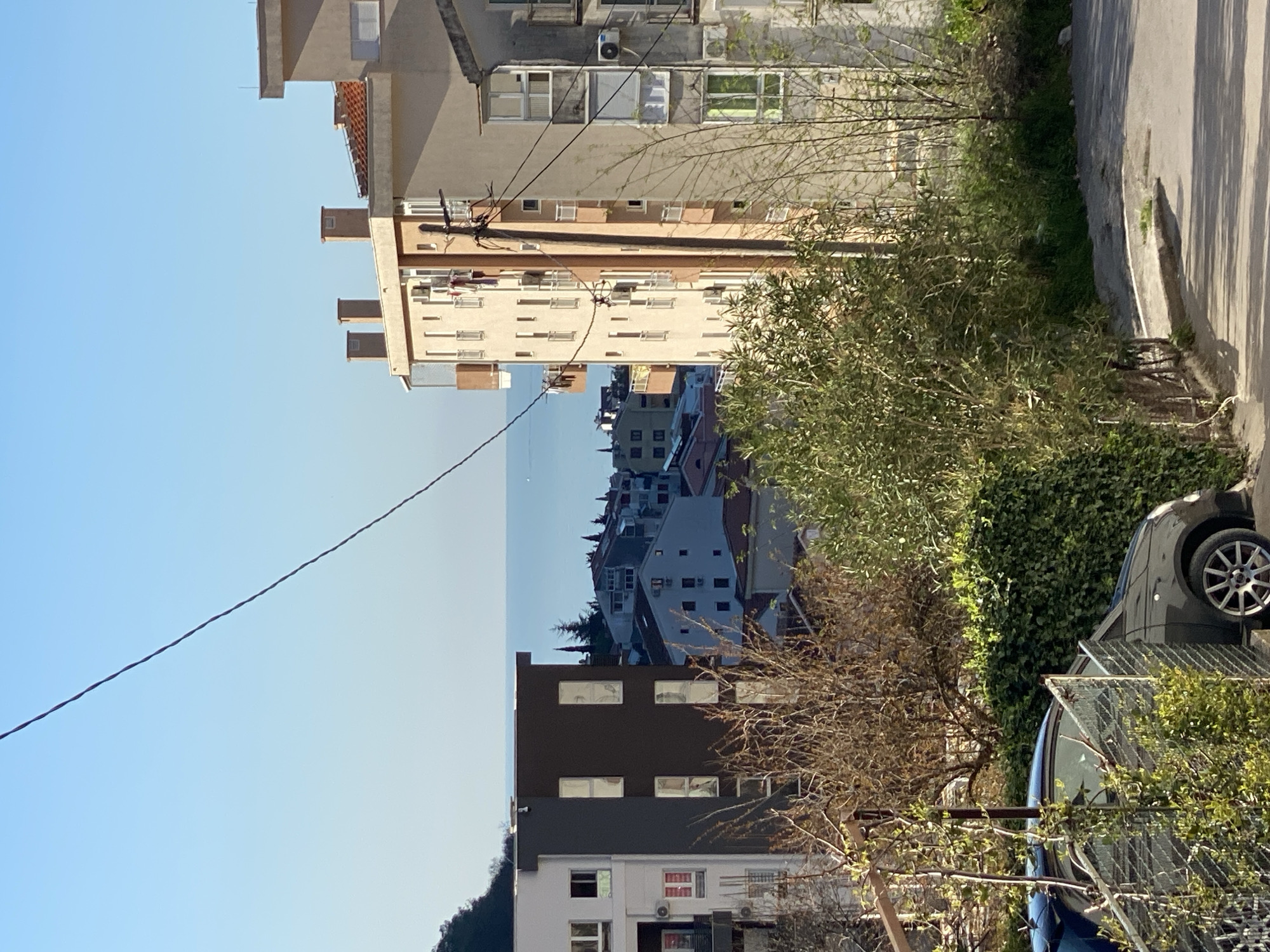 The image depicts a view in Budva, Montenegro, from an elevated position. The skyline is visible between apartment buildings. A power line crosses the frame diagonally from the upper left to the middle right. Green foliage and trees are present, partially obscuring cars parked on the roadside. The buildings are a mix of light and dark colors, with some appearing to be older apartment blocks. A road is visible to the right of the image.