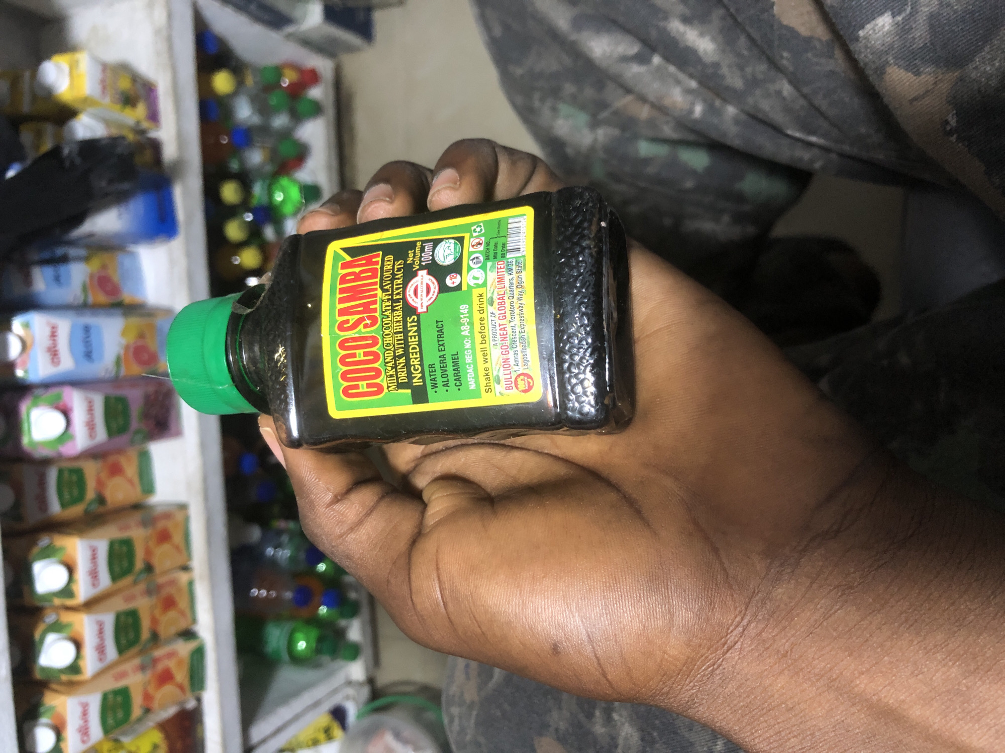 A person's hand holds a small, dark glass bottle with a green cap, labeled "COCO SAMBA MILK AND CHOCOLATE FLAVOURED DRINK WITH HERBAL EXTRACTS." The bottle's label also lists ingredients, a NAFDAC registration number, and contact information for "BULLION GO-NEAT GLOBAL LIMITED" in Lagos. In the background, shelves are stocked with various products, including juice boxes and bottles, suggesting a retail environment like a shop or market. The person's hand and arm are visible, resting on what appears to be camouflage fabric, possibly clothing or a bag. The lighting is moderate, and the image is focused on the bottle and the hand. The scene is likely indoors, in a commercial setting.