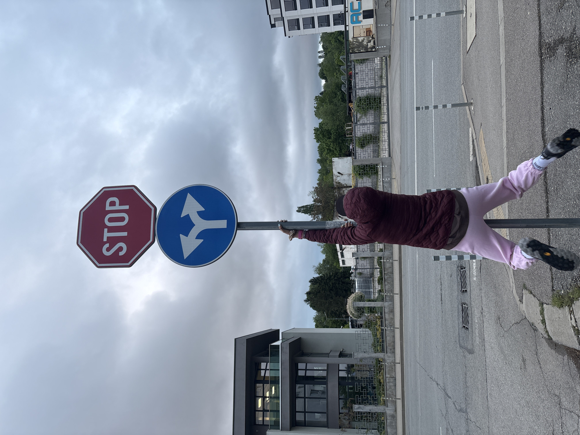 A person wearing a burgundy jacket and light pink pants is hugging a signpost in an urban setting. The signpost has two traffic signs: a red octagonal STOP sign and a blue circular sign with a white arrow splitting in two directions. The background includes buildings, trees, and a cloudy sky. The location appears to be a street corner in the city of Unknown, Unknown.