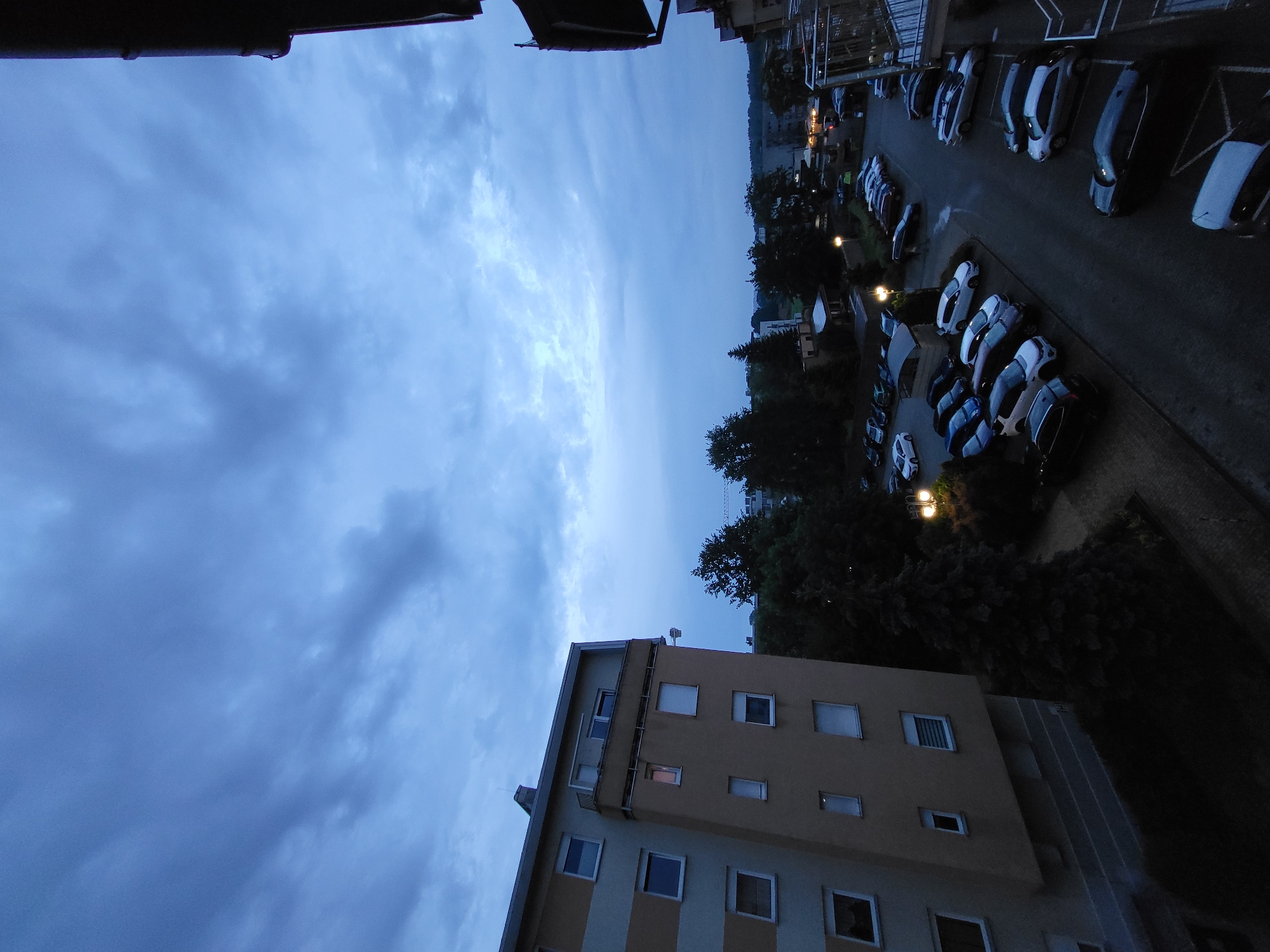 The image captures an evening view from a high vantage point, likely a balcony or window, looking down onto a residential or urban area. The perspective is tilted, making the sky appear on the left and the ground on the right.
The sky, dominating a large portion of the frame, is a deep blue-grey and heavily overcast with dramatic, textured clouds. The light suggests either dusk or early night.
In the foreground, partially cropped, is a multi-story building with a light yellowish-beige facade and multiple windows with light frames. Some windows show faint light from within. A dark, possibly metallic, edge of a roof or balcony overhang is visible across the top of the image.
Below, stretching into the distance, is a street lined with numerous parked cars. Many of these vehicles are modern, with sleek designs, in colors like white, grey, and black. They are arranged in parking bays marked by white lines on asphalt surfaces. Several streetlights are illuminated, casting warm yellow-orange glows onto the street and parking areas.
Dark silhouettes of trees and dense foliage are visible along the street and between buildings, contrasting with the lighter elements and streetlights. Other buildings can be seen in the background, dimly lit.
No people are visible. The scene conveys a quiet, perhaps slightly somber, atmosphere typical of an urban area after sunset. There is no visible text.