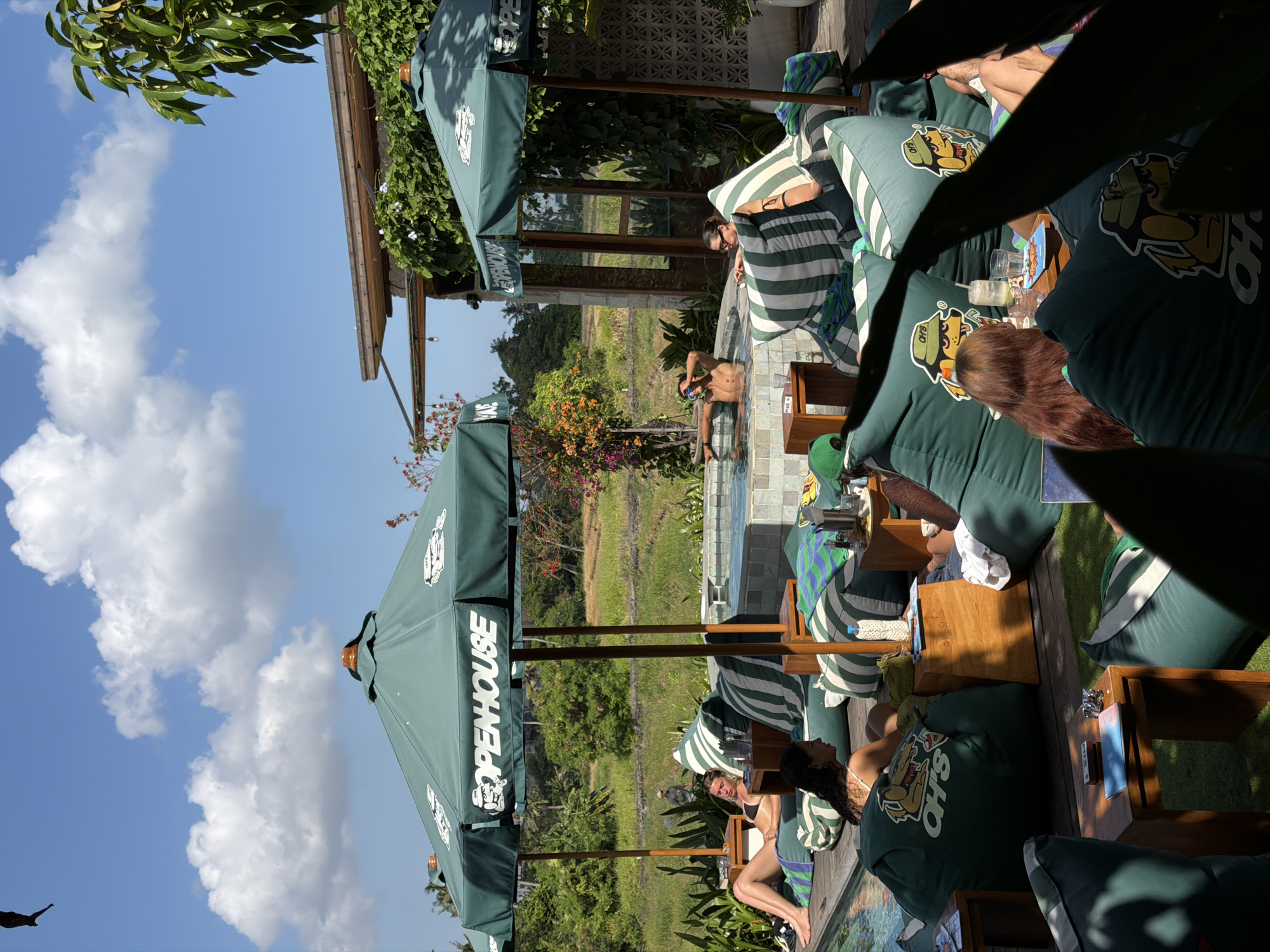 In Mengwi, Indonesia, this outdoor scene depicts a relaxing day at a resort or cafe with a pool and hot tub. Several people are lounging on large green beanbag chairs scattered around a wooden deck and a pool area. Two green umbrellas with "OPENHOUSE" branding provide shade. In the background, lush greenery, including rice paddies and trees, suggests a rural or semi-rural setting. A man is in the hot tub, and a woman is sunbathing by the pool. Others are seated on the beanbags, some with drinks, appearing engaged in conversation or relaxation. The environment is bright and sunny, with blue skies and scattered clouds.