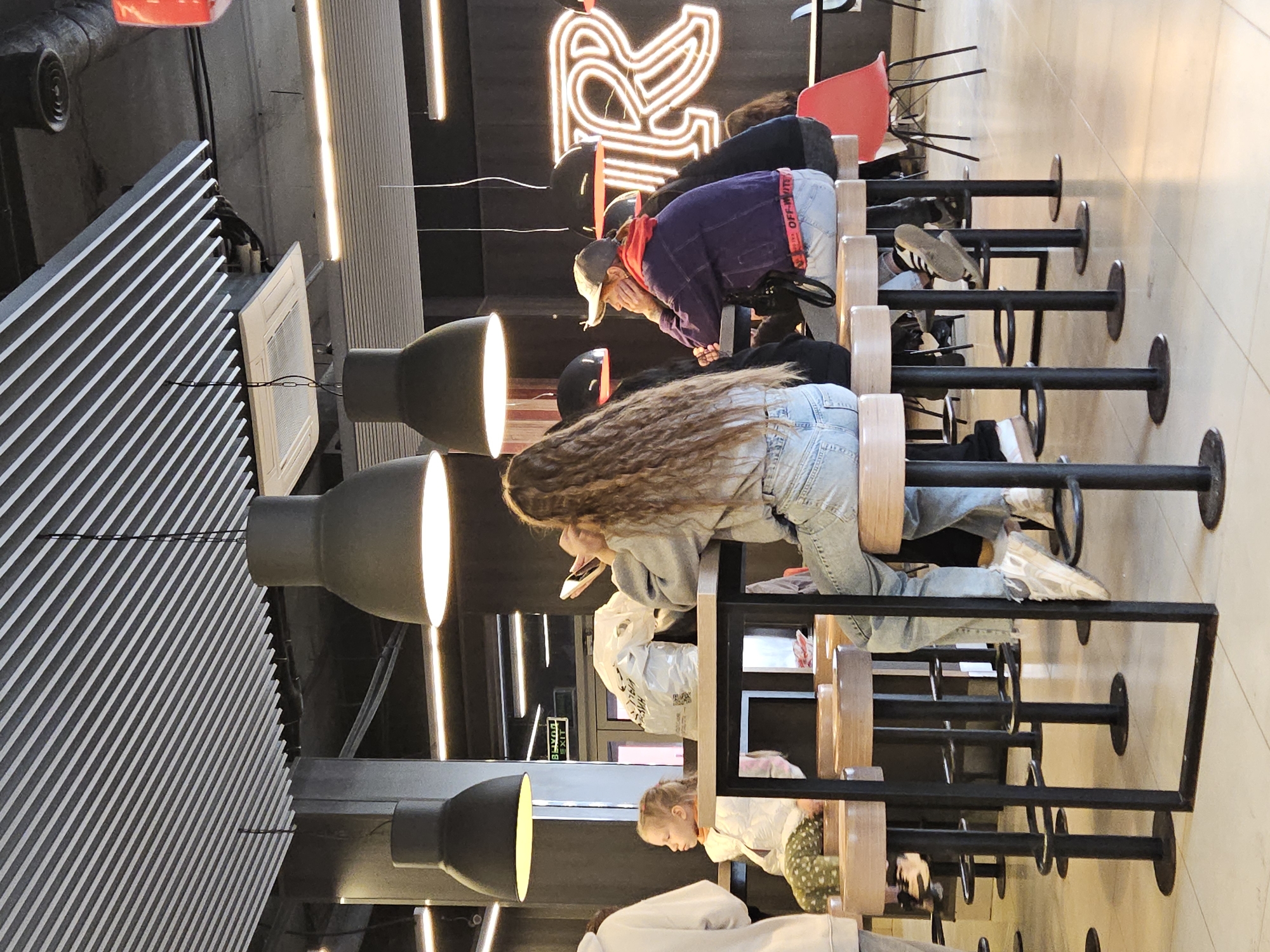 The image depicts the interior of a modern fast-food restaurant, likely a McDonald's given the neon "M" sign. The scene is set in Ulyanovsk, Russia. Several patrons are seated at high-top tables and bar stools. In the foreground, a young woman with long, wavy brown hair is seated at a table, looking at her phone. To her left, a young girl in a white puffer jacket and patterned pants sits on a stool, also appearing to be looking at something at the table. Further to the right, a person wearing a grey baseball cap, purple jacket, and light-colored pants sits with their hands resting on the table. Behind them, other individuals are partially visible. The restaurant features a contemporary design with slatted ceilings, large pendant lights, and neon signage. The floor is tiled, and the overall ambiance suggests a casual dining environment.