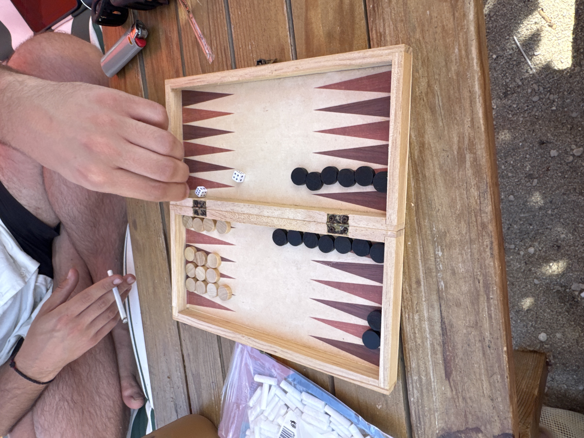 A close-up, overhead view shows a backgammon board laid out on a wooden table outdoors. Two dice, one showing a 5 and the other a 2, are positioned on the board, near two black checkers. To the left, a pile of white backgammon pieces is contained within a clear plastic bag. A person's hands are visible, one holding a lit cigarette, and the other resting near the dice. The background suggests an outdoor setting, possibly a patio or garden, with sandy ground visible below the table. The location is the city of Pallini Municipal Unit, Greece.