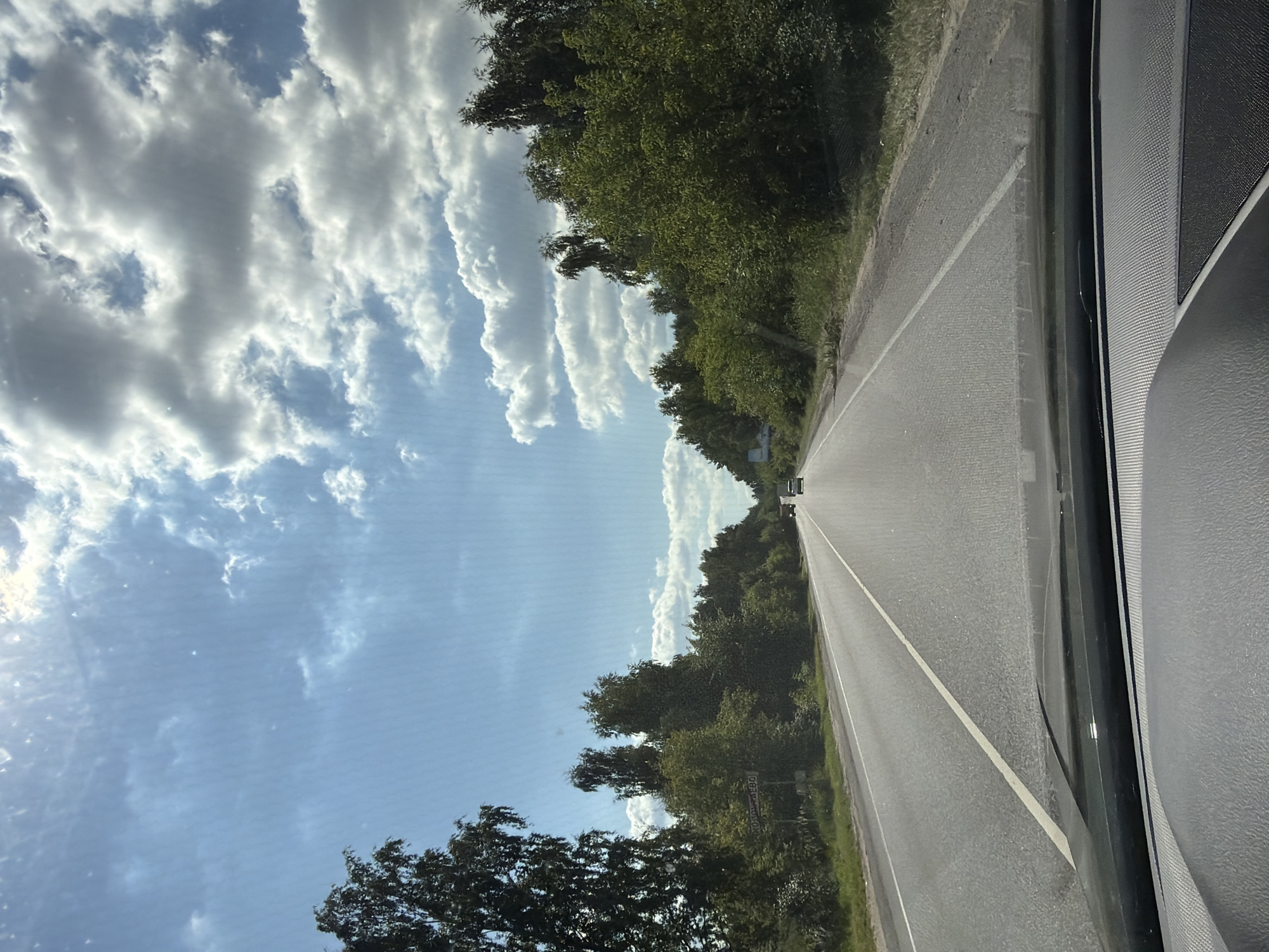 The image captures a view from inside a vehicle driving on a paved road lined with trees and vegetation. The road has two lanes marked by white lines. In the distance, two cars are visible, one white and one darker. The sky above is blue with scattered white clouds. On the left side of the road, a sign with Cyrillic text is partially visible, indicating "КОЛОНТАЕВО" (Kolontaevo). The foreground shows the dashboard of the vehicle. The scene appears to be a rural or suburban area in Kolontaevo, Russia, during daylight.