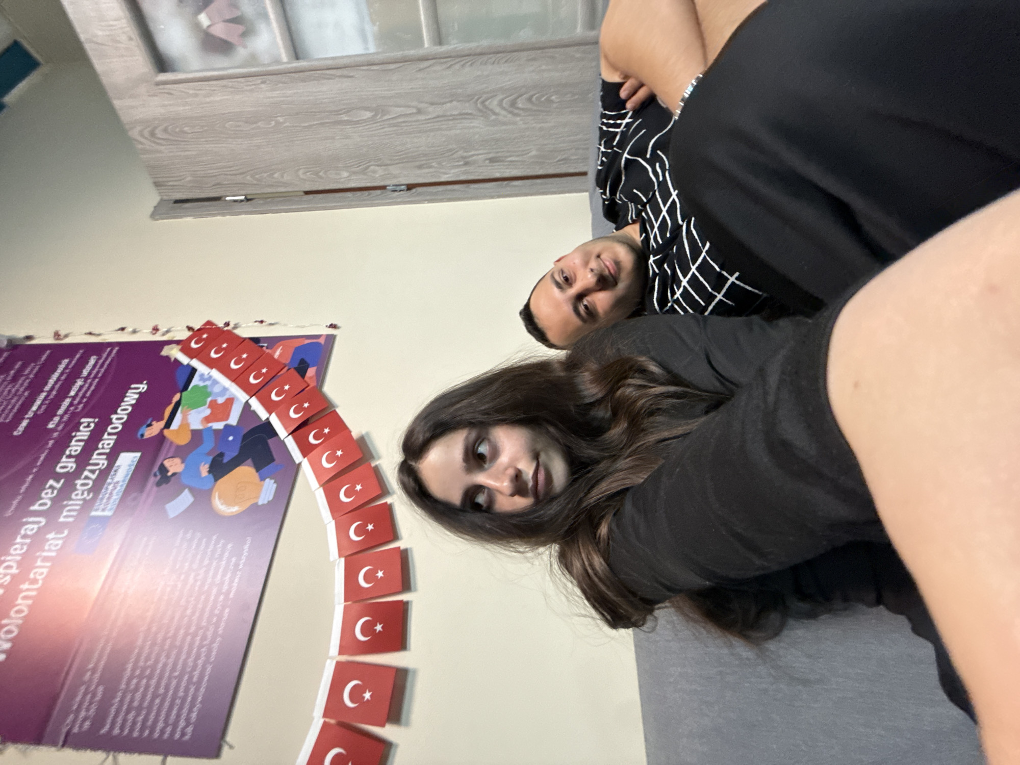 The image captures two young individuals, a male and a female, relaxing indoors, possibly on a sofa or couch. The scene is rotated 90 degrees counter-clockwise from its natural orientation.
When viewed correctly (rotated clockwise), a young woman with long, dark brown hair is in the foreground, lying on her side and looking towards the camera with a neutral, slightly curious expression. She is wearing a black top. Behind her, a young man with short dark hair and a beard is also lying down, smiling faintly and looking directly at the camera. He is wearing a black shirt with a white grid pattern. Both appear relaxed and comfortable.
The setting is an indoor space, likely a living room or common area, with a light-colored wall. To the left of the individuals, there is a large, reddish-purple poster affixed to the wall. The poster features illustrations of people and contains Polish text. Below the poster, and arching around its edge, is a string of small, rectangular flags, each depicting the red background with a white star and crescent moon, which is the national flag of Turkey. Above the poster, a light wooden panel or door frame is visible.
Visible text on the poster, identified through OCR, includes phrases such as "wspieraj bez granic!" (support without borders!), "wolontariat międzynarodowy." (international volunteering.), and "Czas trwania mobilności" (Duration of mobility). These phrases, combined with the presence of Turkish flags and the location context of Opole, Poland, suggest the scene might be related to an international youth exchange, volunteering program, or a cultural event. The overall mood is casual and friendly.