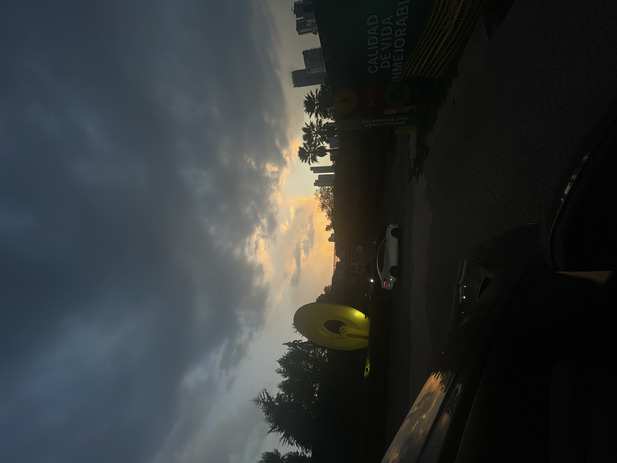 The image is taken at dusk in what appears to be Huixquilucan de Degollado, Mexico. The photograph, likely taken from inside a vehicle, looks out toward a large yellow circular sculpture set against a backdrop of trees. Behind the trees, the sky is a blend of grey storm clouds and a strip of orange sunset. In the distance, modern high-rise buildings are visible. To the right, there's a building or sign that reads "Calidad de Vida Inmejorable". A white car is visible further down the road. Part of the car's interior is visible on the right side of the image.