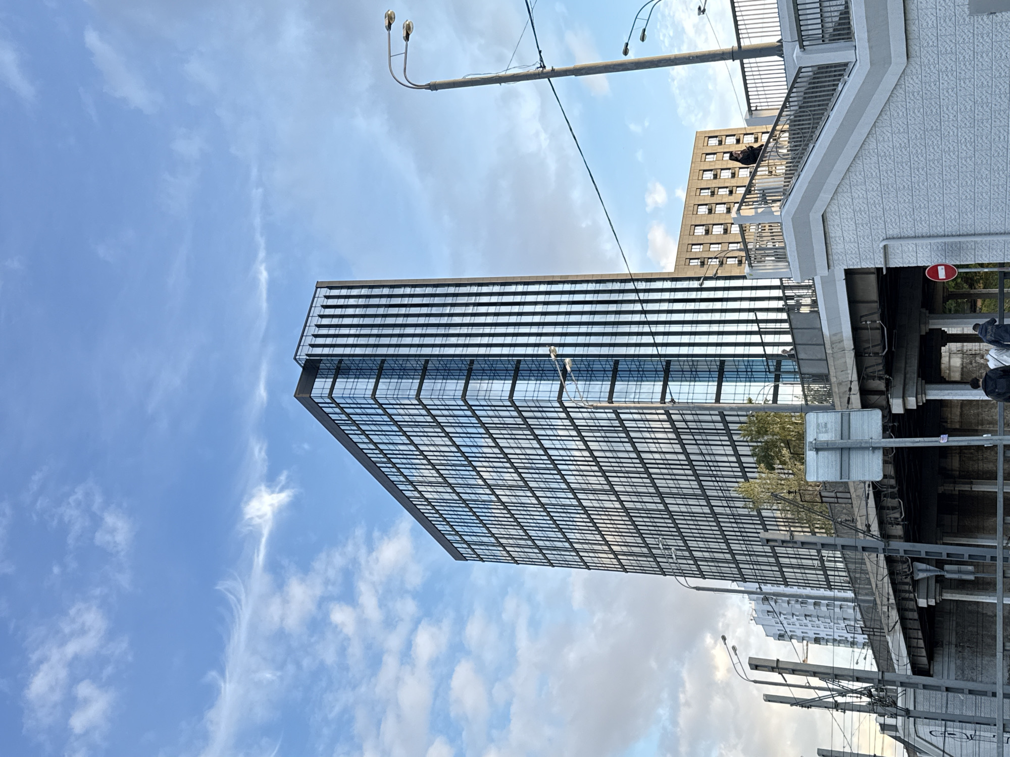 The image depicts an urban street scene in Moscow, Russia, viewed from below and rotated, showing a mix of modern and older architecture under a bright, partly cloudy sky.
In the center-left, a prominent modern skyscraper with a reflective glass facade rises vertically, its numerous windows reflecting the blue sky and scattered white clouds. To the right and slightly behind this building, another tall, light-colored building with many rectangular windows is visible. An elevated structure, possibly a bridge or overpass with a metal railing, stretches horizontally across the right side of the frame, constructed of white, textured material. Below this structure, various urban elements like support pillars, other buildings in the background, and industrial-looking infrastructure are visible.
A long street light pole with multiple lamp heads extends diagonally into the sky from the upper right, and several utility wires crisscross the frame. A red circular traffic sign with a white horizontal bar (indicating "No Entry") is partially visible beneath the elevated structure on the right. Another rectangular gray street sign is also present, but its text is not decipherable. Green foliage from trees can be seen peeking out from behind the elevated structure and near the base of the glass building.
The scene is static, devoid of explicit human activity, suggesting a typical daytime view of a bustling city. The lighting indicates it is daytime, likely late morning or afternoon, with clear weather conditions featuring a bright blue sky and scattered white and wispy clouds.