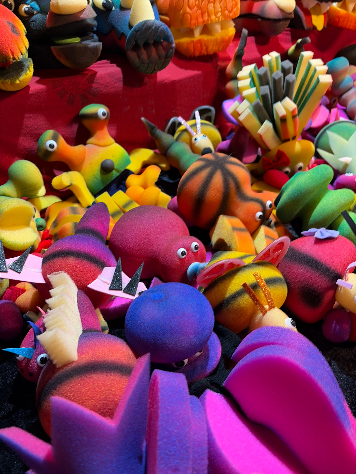 The image displays a vibrant collection of handcrafted felt figurines, possibly for sale or as decorative items, arranged on a red cloth surface. The figurines are diverse in shape and color, featuring cartoonish eyes and simple designs that suggest characters from children's media or imaginative creatures. Notable among them are bee-like figures with yellow and black stripes, round red and purple creatures, and other abstract shapes in various hues like green, orange, and yellow. Some figures have exaggerated features such as spikes or antennae. In the background, other colorful items are visible, contributing to a festive and playful atmosphere. The setting does not provide specific clues about its location in Salou, Spain, or any particular activity beyond the display of these objects. The focus is entirely on the handmade figures and their arrangement.