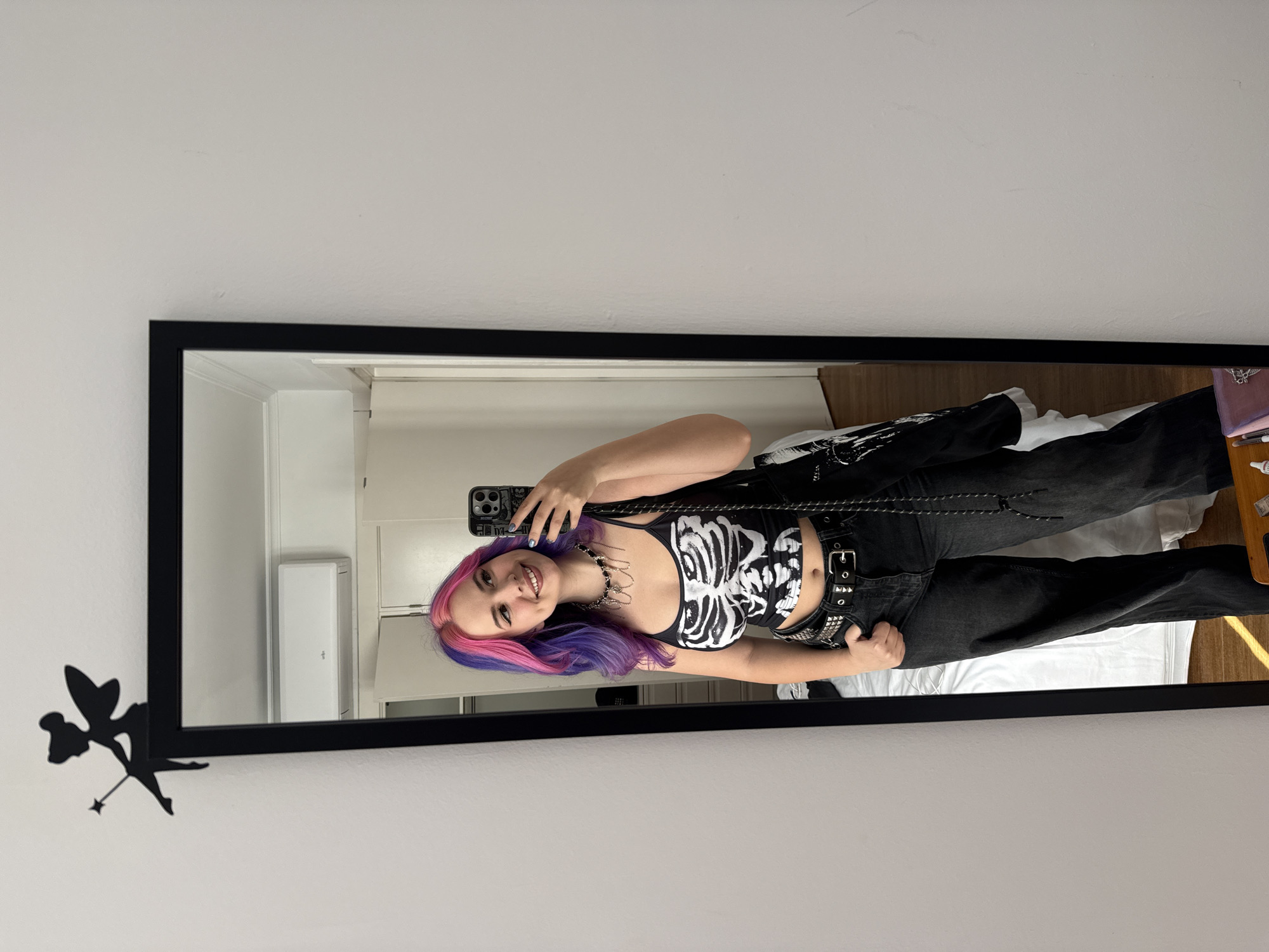 A young woman with pink and purple hair is posing for a picture in front of a full-length mirror. She is wearing a black and white skeleton-print top, black pants with a studded belt, and a chain necklace. She is holding her phone up to take a selfie. The mirror has a black frame and a small fairy silhouette attached to the bottom left corner. The reflection shows a room with a white air conditioning unit on the wall. The bed is covered with white sheets and a black jacket, and there's a pink purse and some small items on a wooden surface next to it. The location is likely a bedroom or apartment, but there is nothing in the image to indicate that the location is in the city of Madrid, Spain.