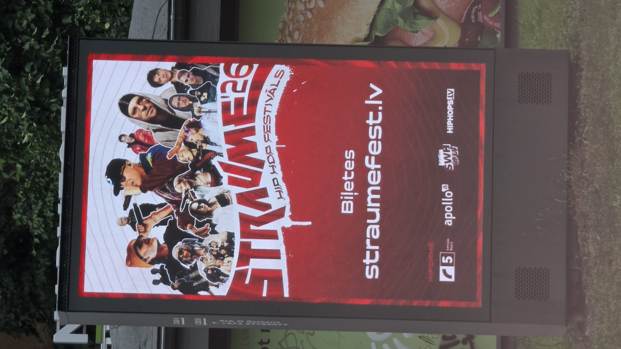 This is a photograph of an outdoor digital billboard displaying an advertisement for a hip-hop festival. The billboard is oriented vertically and is situated outdoors, likely in an urban environment given the presence of grass and foliage in the background. The advertisement features a collage of images of people, predominantly young individuals, some with microphones and in what appear to be performance poses, suggesting a music event. The background of the advertisement is a bright red with white concentric circles emanating from the center.
Prominently displayed in large, stylized white font is the word "STRAUME" followed by "26" and "HIP HOP FESTIVALS". To the right of this main text, in white, is the word "Biļetes" (Tickets in Latvian) and then "StraumeFest.lv", which is likely the event's website. Below this, several logos are visible: "INFORME", a "75" logo, "apollo", a logo that appears to be "SWH" or "SWHJ", and "HIPHOPSLV".
The overall mood conveyed by the advertisement is energetic and youthful, consistent with a hip-hop festival. The lighting suggests it is daytime. There are no clear indications of weather conditions, but the image is clear and sharp. The location cues, such as the potential presence of Latvian text and logos, combined with the provided context, point to Riga, Latvia as the location.