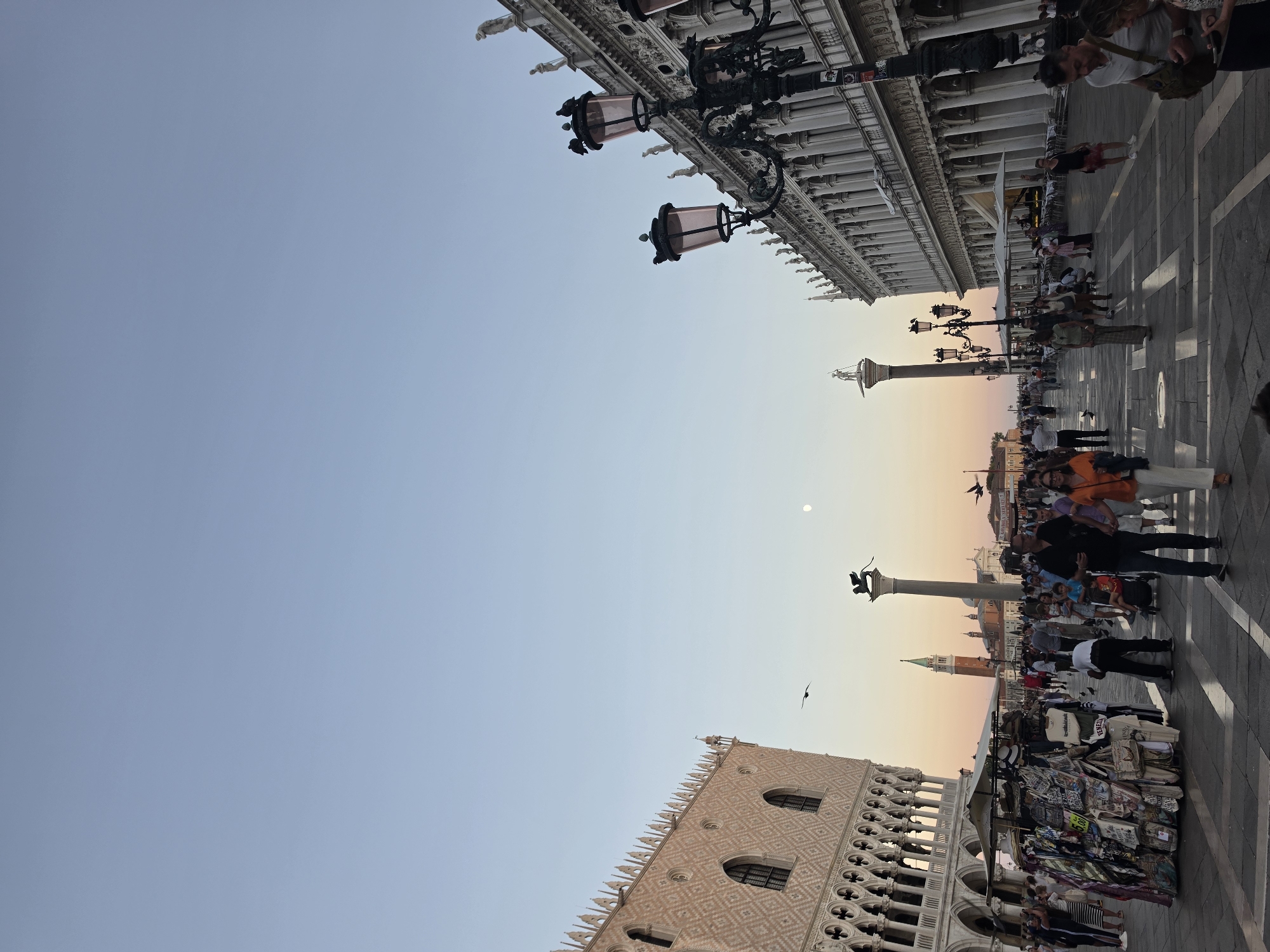 This image depicts St. Mark's Square (Piazza San Marco) in Venice, Italy, during what appears to be late afternoon or early evening, given the warm light and clear sky. In the foreground, a bustling crowd navigates the paved square. A kiosk selling souvenirs, primarily t-shirts and bags, is positioned on the left side of the image. Numerous people are walking, some holding hands, others with children, and some are gathered around the kiosk. Two ornate lampposts with multiple lights stand prominently on the right, framing a grand building with a colonnaded facade. On the left, a large, intricately decorated building, likely the Doge's Palace, is partially visible. In the middle ground, two tall columns stand, topped with statues, with a bell tower in the distance. A faint moon is visible in the sky. The overall atmosphere is that of a popular tourist destination with significant pedestrian traffic.