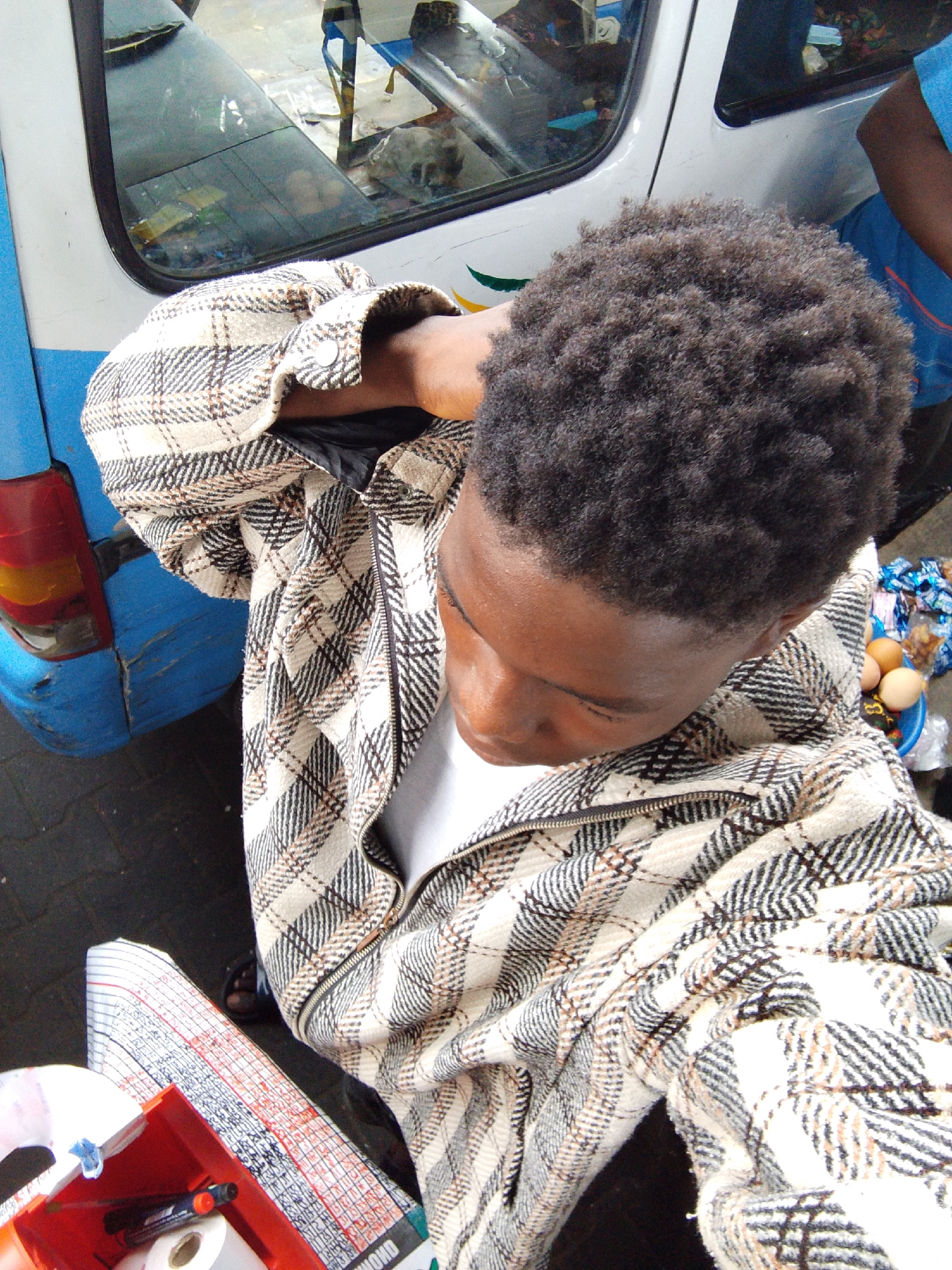 An overhead shot captures a young Black person in an outdoor, urban setting in Lekki, Nigeria. The person, likely male, is the central subject, looking downwards and slightly to their right. They have short, dark, tightly curled hair and wear a beige, black, and brown plaid zip-up jacket over a white t-shirt. Their right arm is raised, possibly holding the camera for a selfie or adjusting their clothing.
Behind the subject, a blue and white commercial vehicle, possibly a minibus, is visible. The blue rear section shows a red taillight and some scuffs. The white side of the vehicle features a green and yellow decal. Through its window, blurred reflections and interior contents are visible.
To the subject's right, a blue plastic bowl holds several brown eggs and various packaged snacks or candies, suggesting a street vending context. In the foreground, a red plastic tray or box contains a roll of white material (possibly tape or paper) and a few pens or markers. Beneath this tray, a white and red patterned fabric, possibly a table cloth or mat, is spread out, displaying columns of text or numbers in black and red. The word "DHOND" is faintly visible on this fabric. Another person's arm, wearing a blue shirt, is partially visible near the white vehicle in the upper right background. The scene appears to be taking place during the day under natural light.