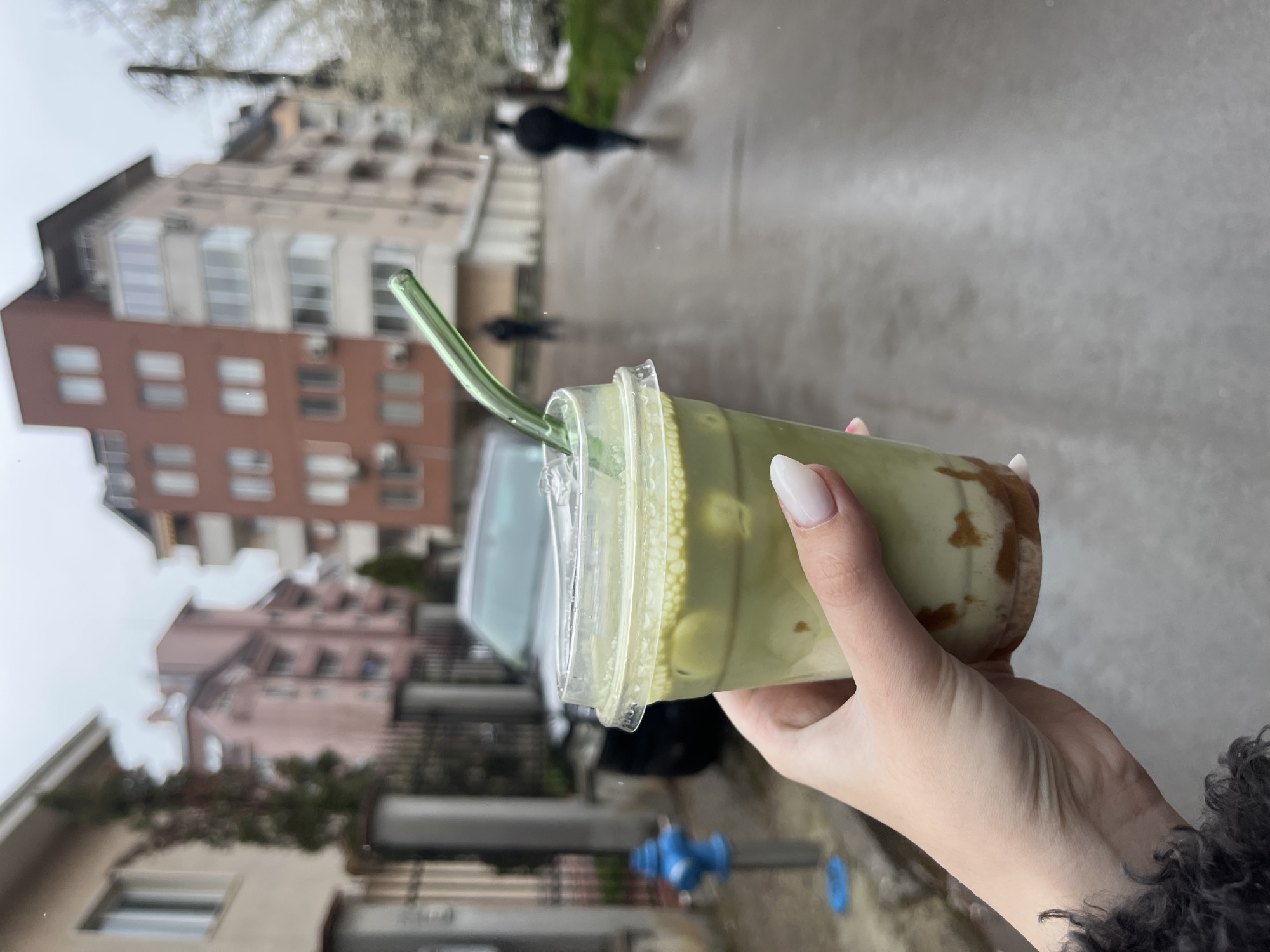 A woman with pale, manicured fingernails is holding a clear plastic cup filled with a green beverage with brown swirls at the bottom. The drink also contains small, white tapioca pearls. A green glass straw is inserted into the drink. The woman is wearing a garment with a textured, possibly fur, collar.
The background reveals an urban setting, likely a street in Sofia, Bulgaria. The road appears wet, possibly from rain. A person is walking in the background. Buildings of varying heights and colors line the street, including a brown brick building and a pink building with visible windows and balconies. A dark-colored car is partially visible. There is a blue fire hydrant on the sidewalk. The sky is overcast and gray, suggesting a cloudy day.