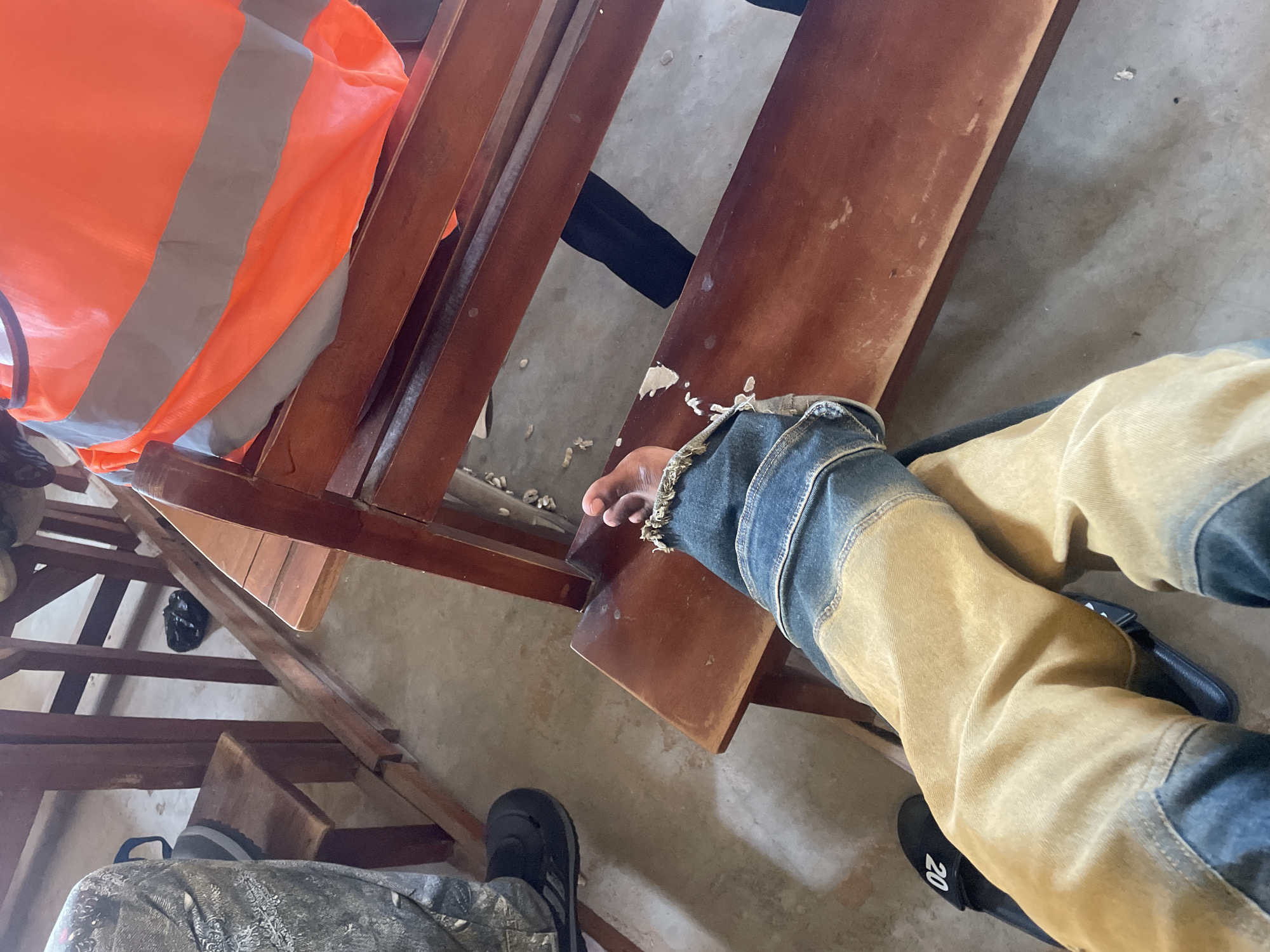 The image shows a person's legs and feet, clad in distressed denim jeans and a tan jacket, resting on wooden benches. The person is wearing black slide sandals with the number "20" visible on the right sandal. A bright orange safety vest with reflective stripes is draped over the wooden structure to the left. The setting appears to be outdoors or in a poorly lit indoor space, with a concrete floor and wooden benches. The overall impression is of a casual or work-related environment. There is some debris on the floor, suggesting a construction or maintenance area. The lighting is dim and appears to be natural daylight, casting shadows on the floor. No specific time of day or weather conditions can be determined. The image does not convey any discernible emotion. The location context of Lokoja, Nigeria, does not provide further visual cues within the image itself.